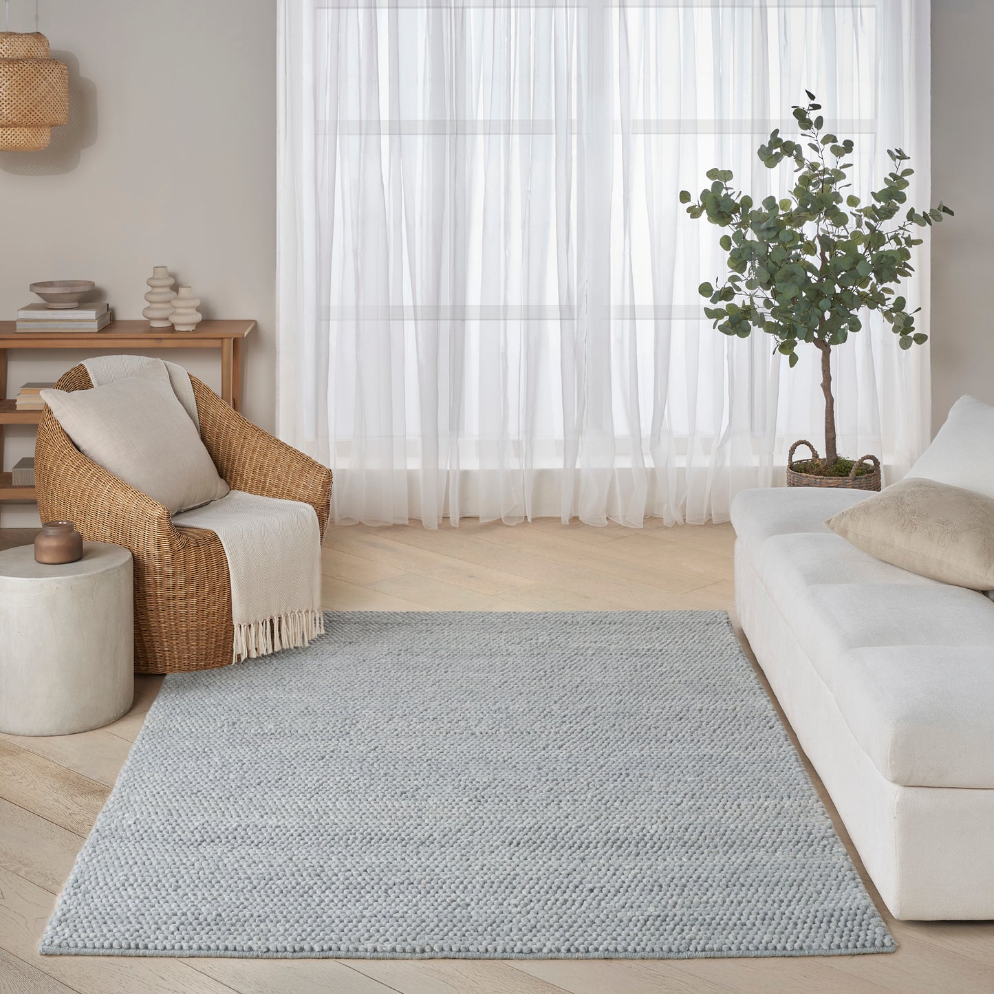 Bright living room with light wood floors, gray woven rug, white sofa, wicker armchair, sheer curtains, & a potted plant.