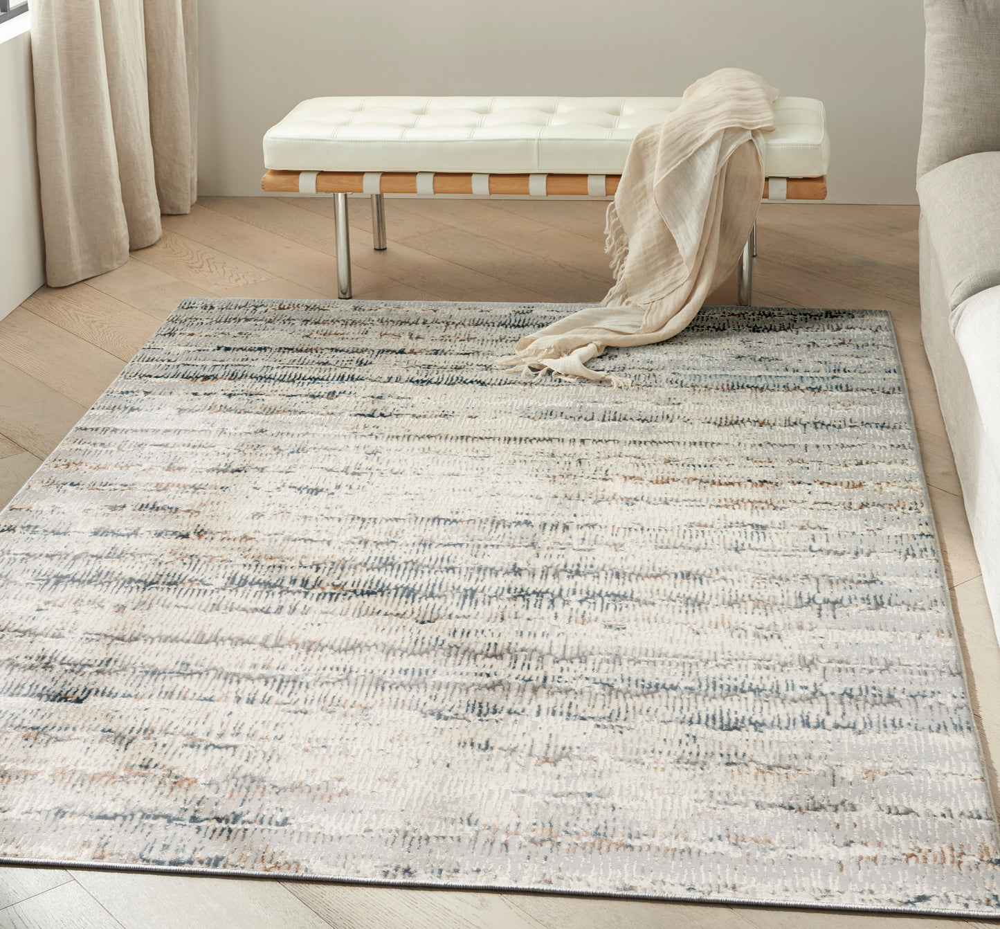 Cream tufted bench with wooden slats and metal legs, draped beige throw, on a gray-blue abstract rug in a bright living room.