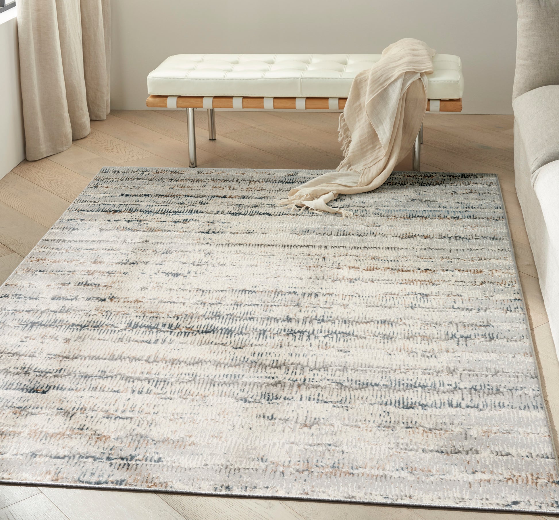 Cream tufted bench with wooden slats and metal legs, draped beige throw, on a gray-blue abstract rug in a bright living room.
