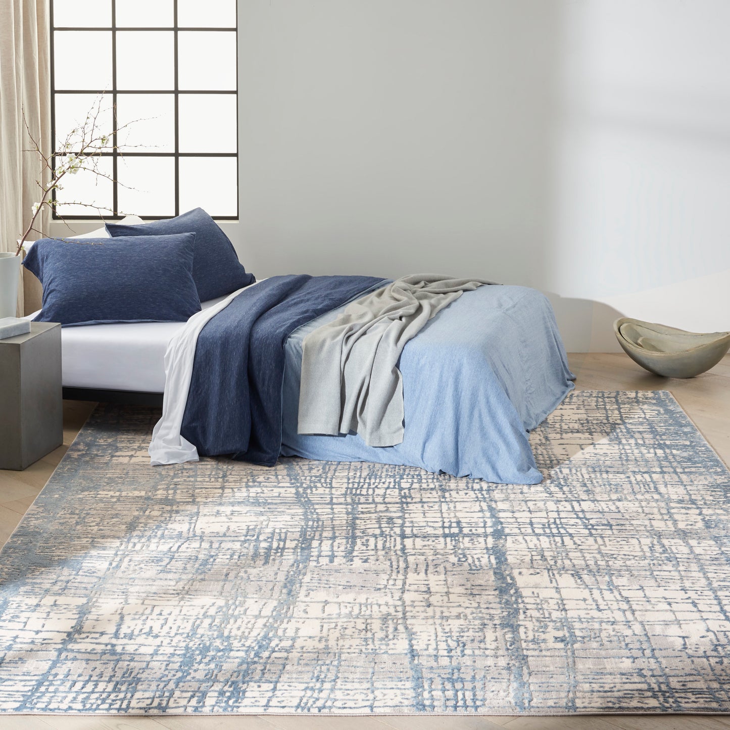Blue bedding with navy and light blue layers, gray throw on bed, over a blue-cream abstract rug in a minimalist bedroom.