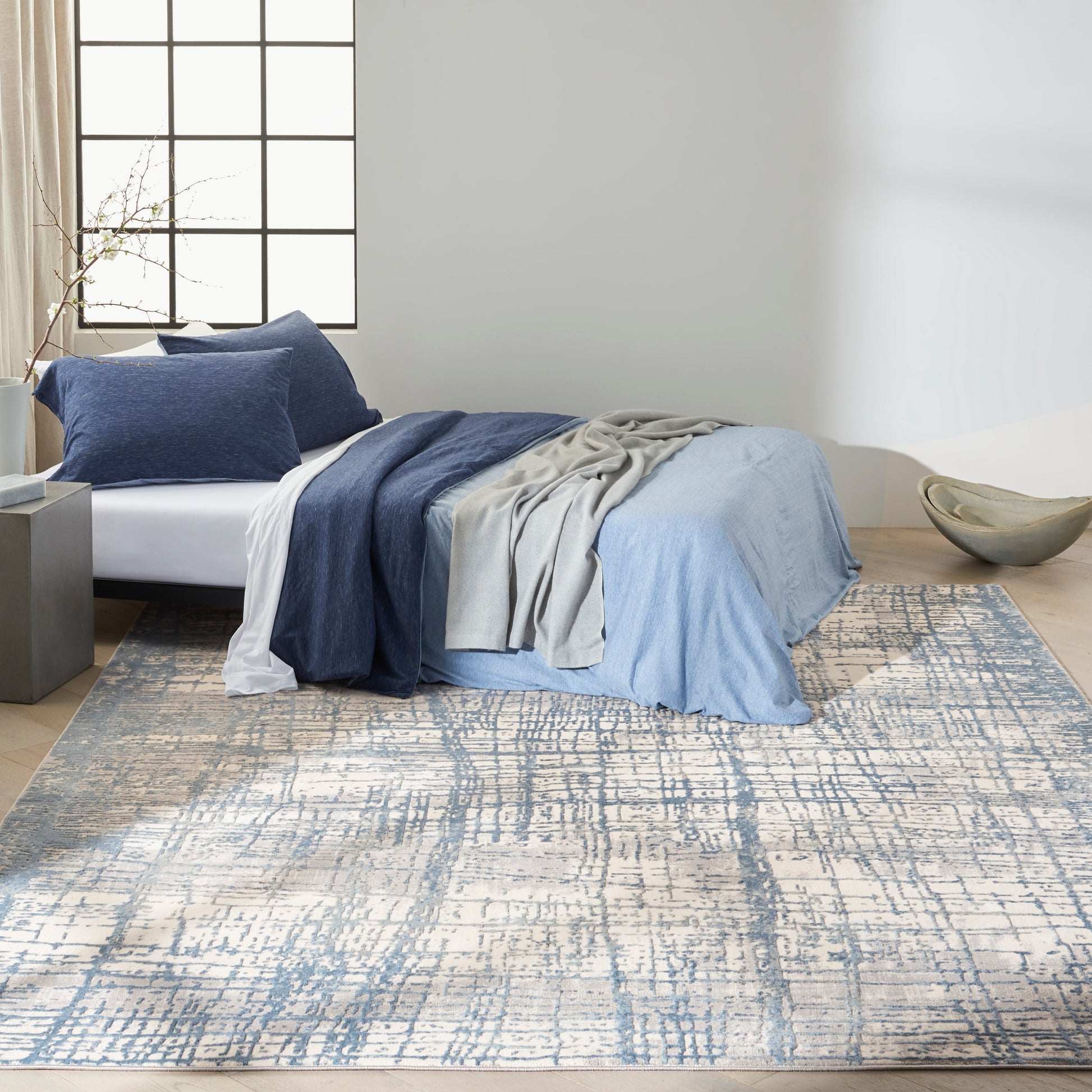 Blue bedding with navy and light blue layers, gray throw on bed, over a blue-cream abstract rug in a minimalist bedroom.
