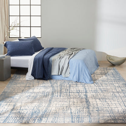 Blue bedding with navy and light blue layers, gray throw on bed, over a blue-cream abstract rug in a minimalist bedroom.