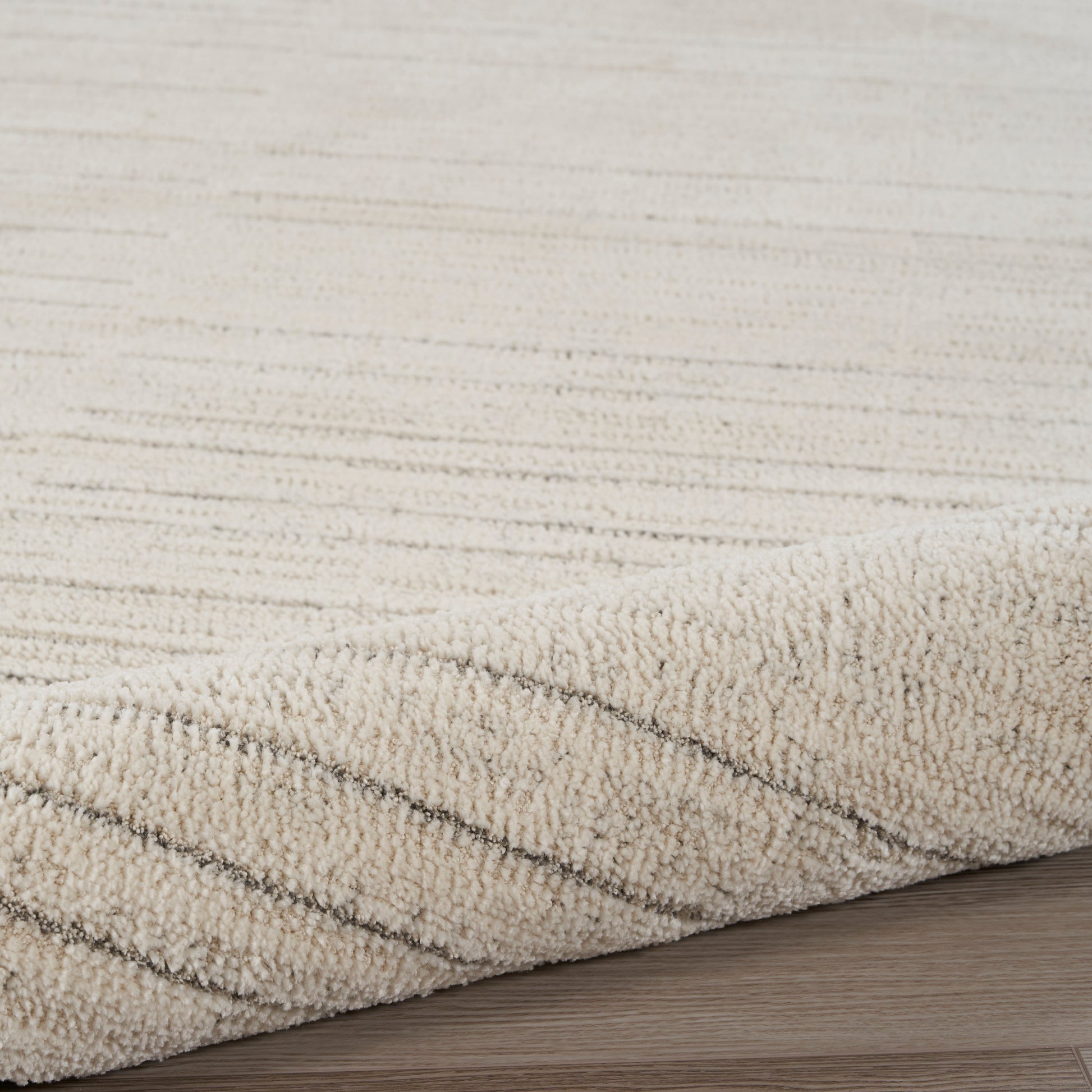 Beige plush rug with subtle diagonal stripes, soft textured pile, edge curled on light wood floor.