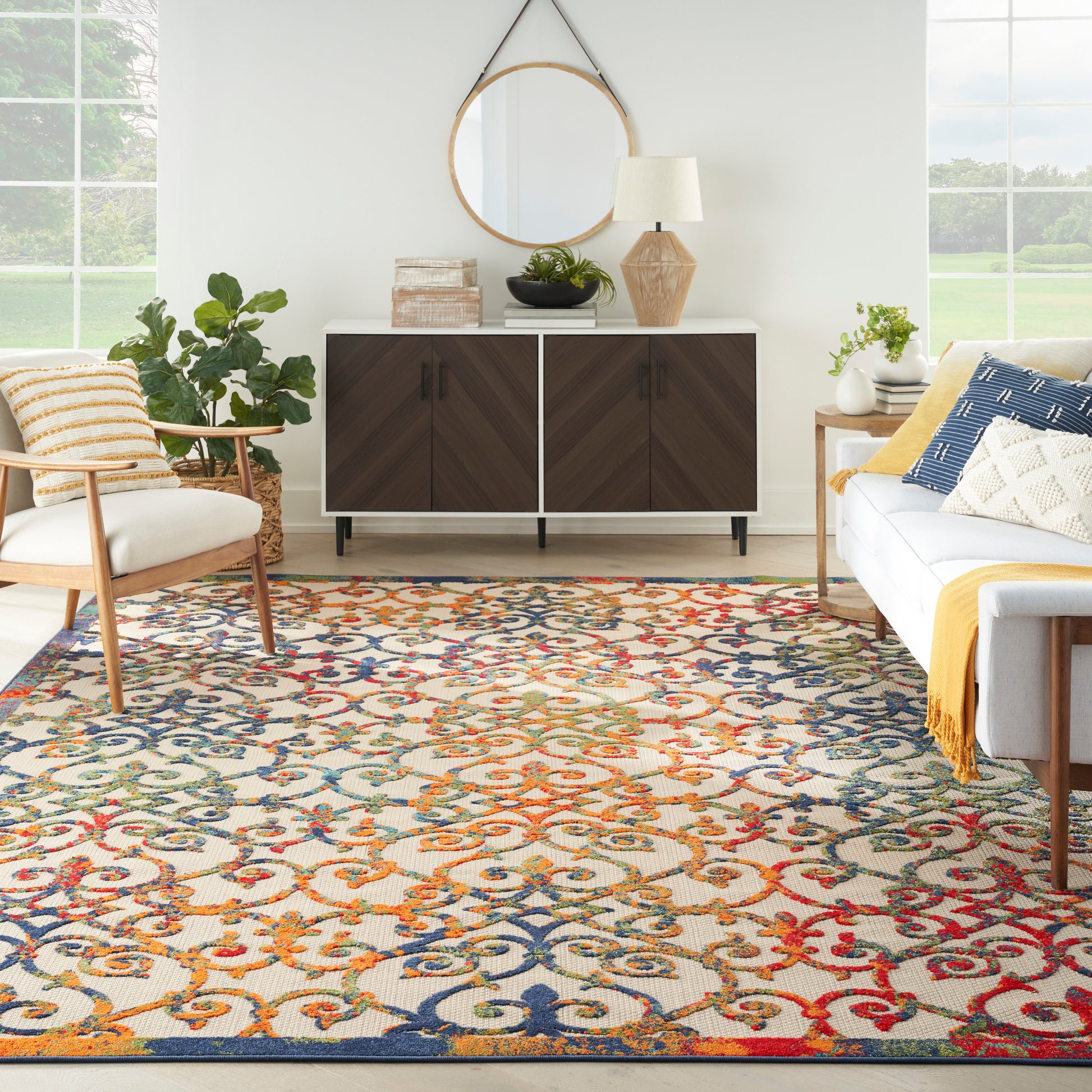 Living room with a vibrant multicolor rug, dark wood sideboard, round mirror, plants, and mid-century seating.