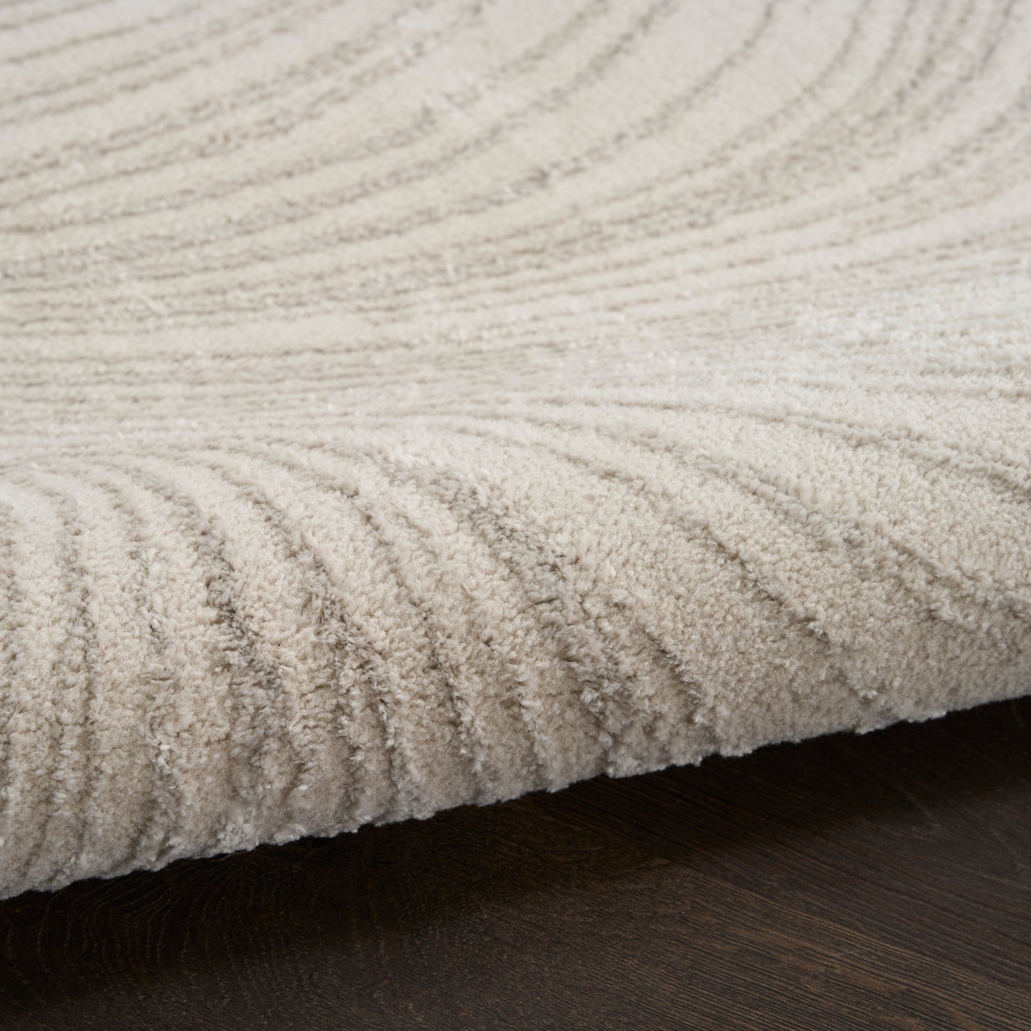 Beige textured pile area rug with concentric wave pattern on dark wood floor.