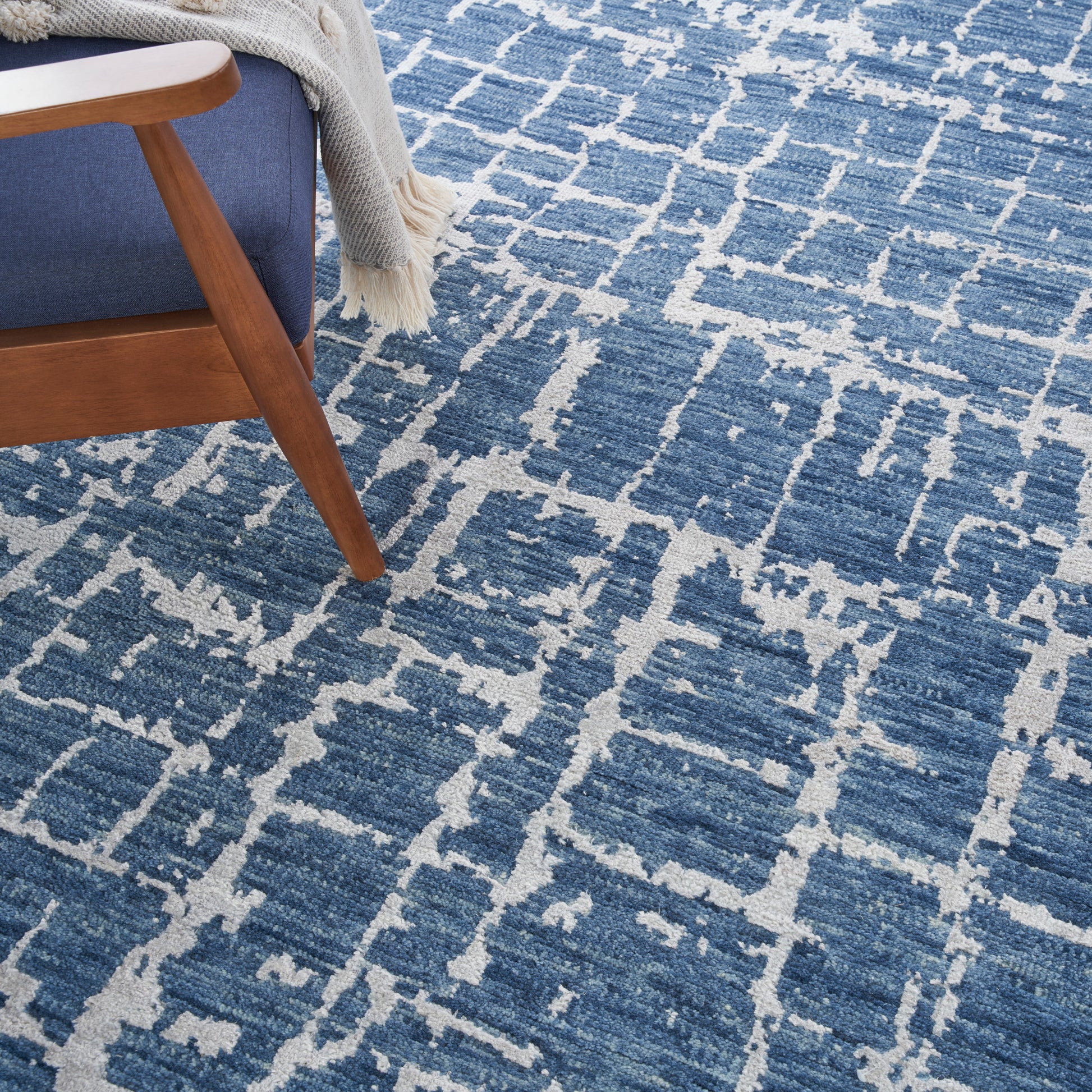 Blue and cream geometric area rug with distressed motif, paired with a mid-century wooden chair and beige knit throw.