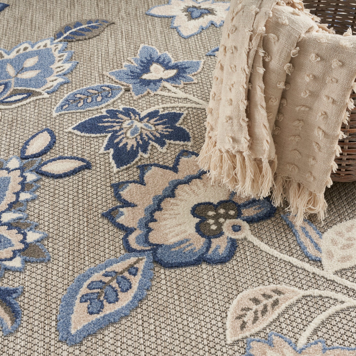 Beige floral area rug with blue and cream blossoms; textured weave, with a fringed beige throw draped over a wicker basket.