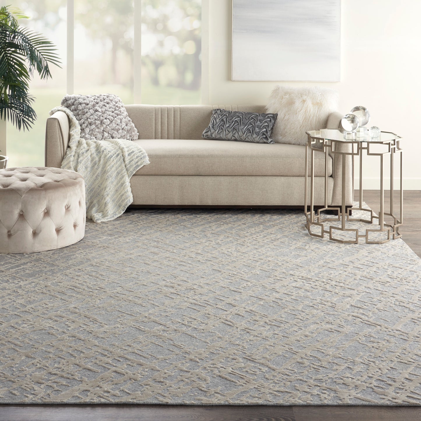 Beige sofa with textured throws and cushions, round tufted ottoman, gold geometric side tables, and a gray patterned rug.