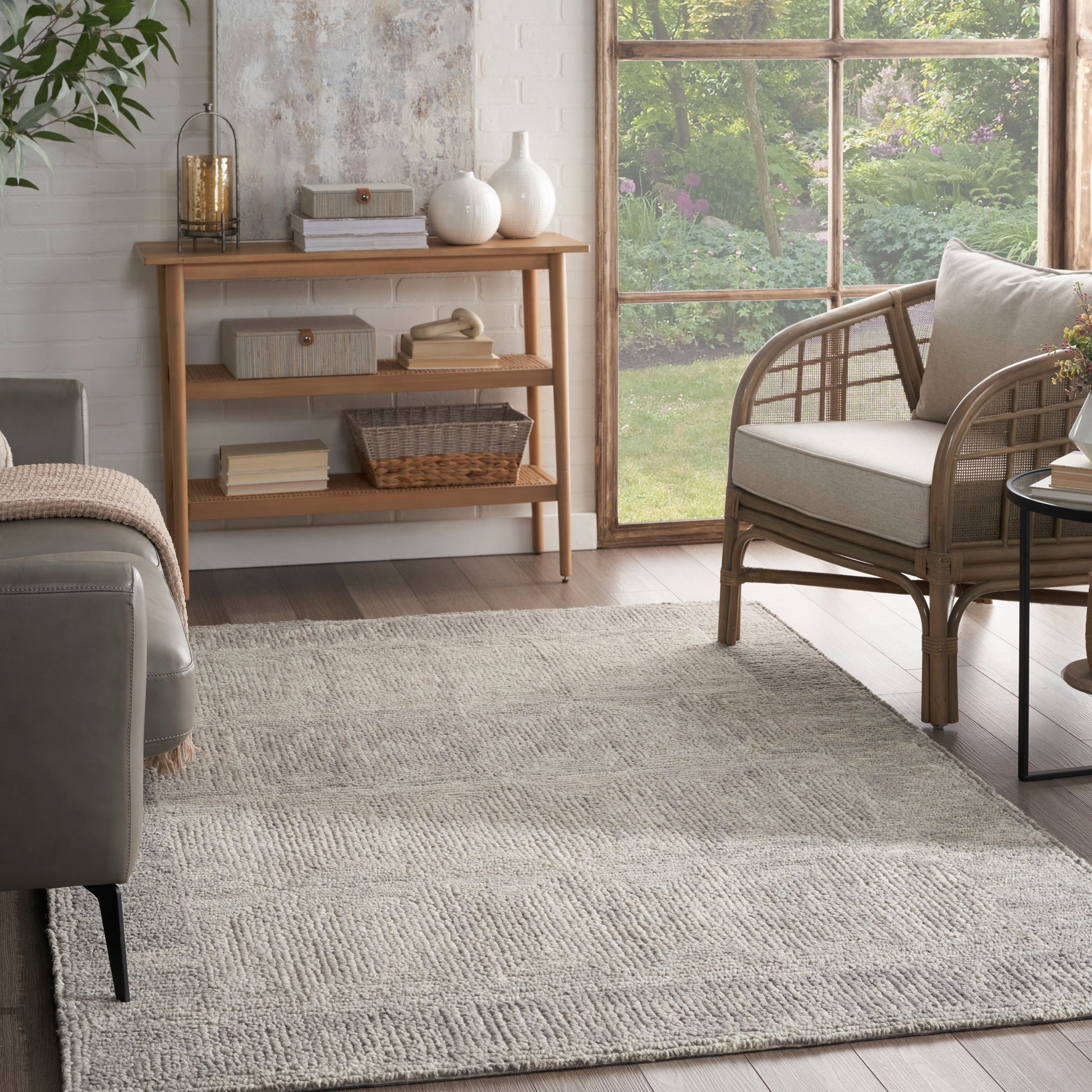 Neutral living room with beige textured rug, light wood shelving, rattan armchair, gray sofa, and garden view.