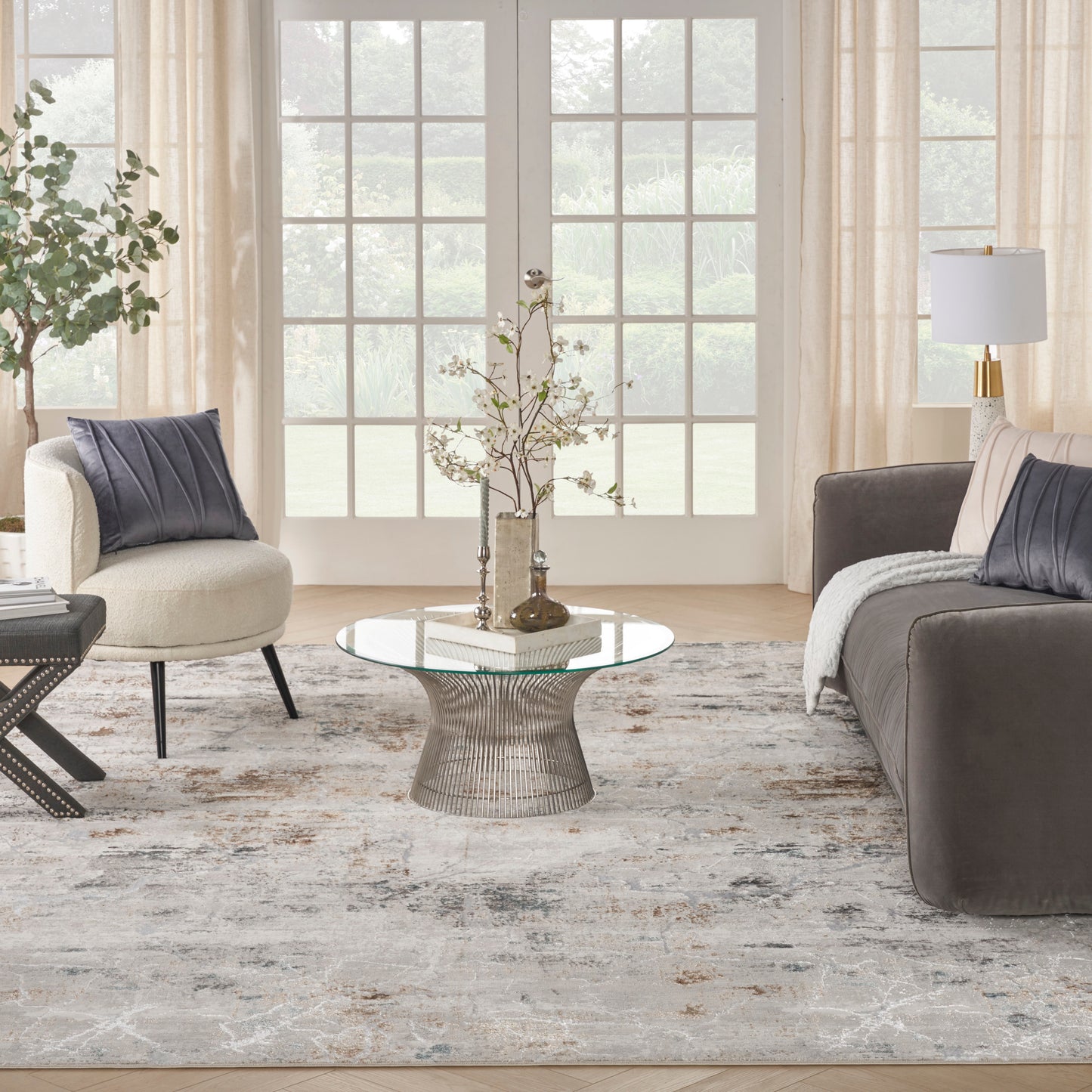 Bright living room with glass doors, cream armchair, gray sofa, round glass coffee table with metal base, neutral rug.