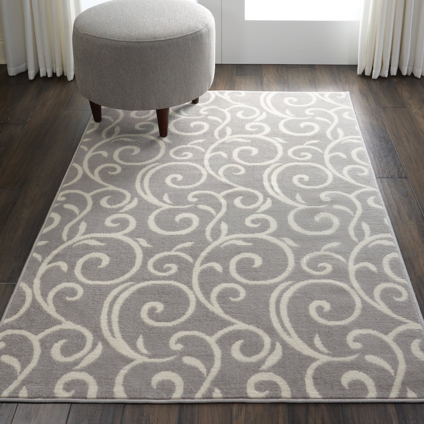Light gray rug with ivory swirling scroll pattern, round gray ottoman with wooden legs on dark wood floor.
