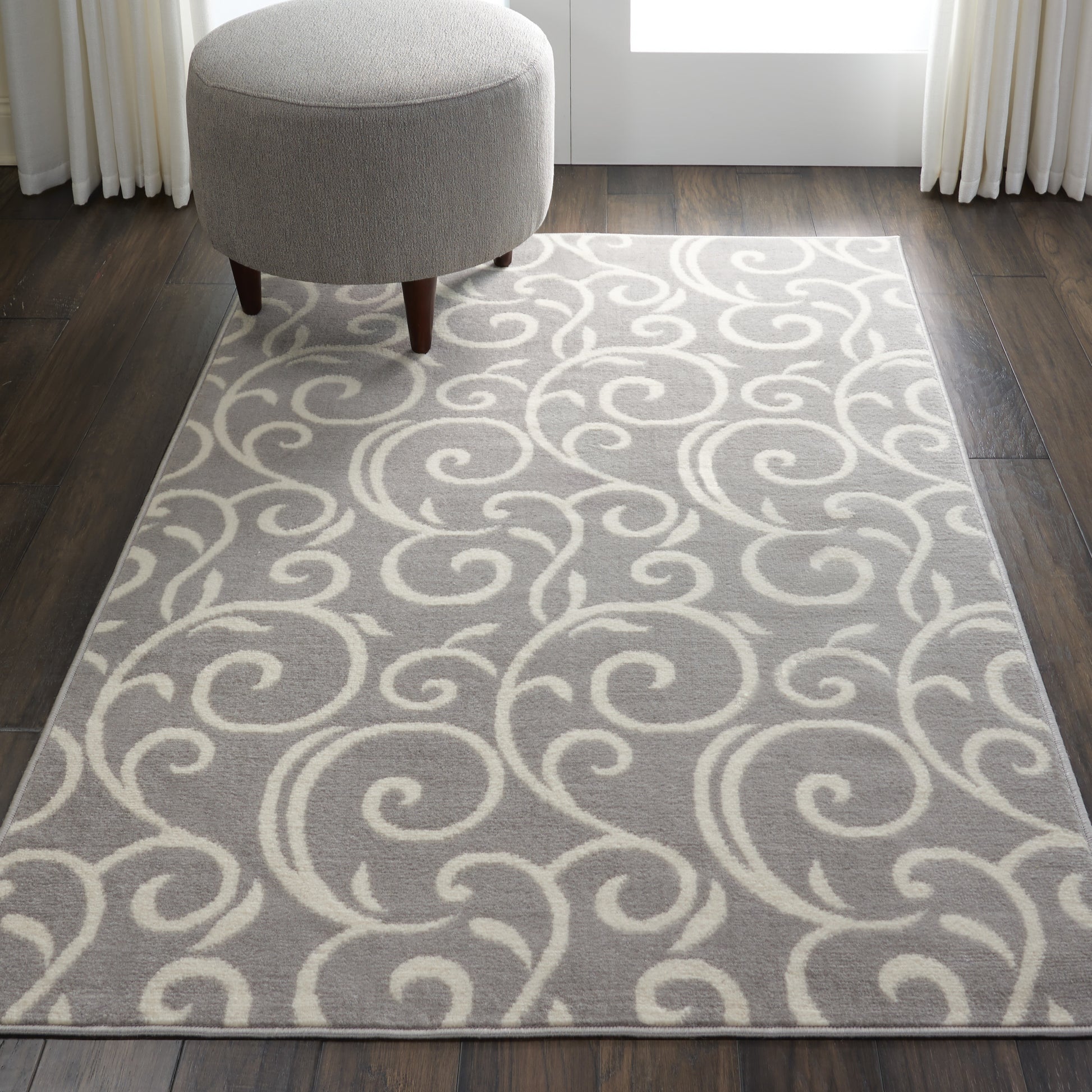 Light gray rug with ivory swirling scroll pattern, round gray ottoman with wooden legs on dark wood floor.