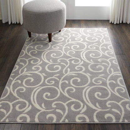 Light gray rug with ivory swirling scroll pattern, round gray ottoman with wooden legs on dark wood floor.