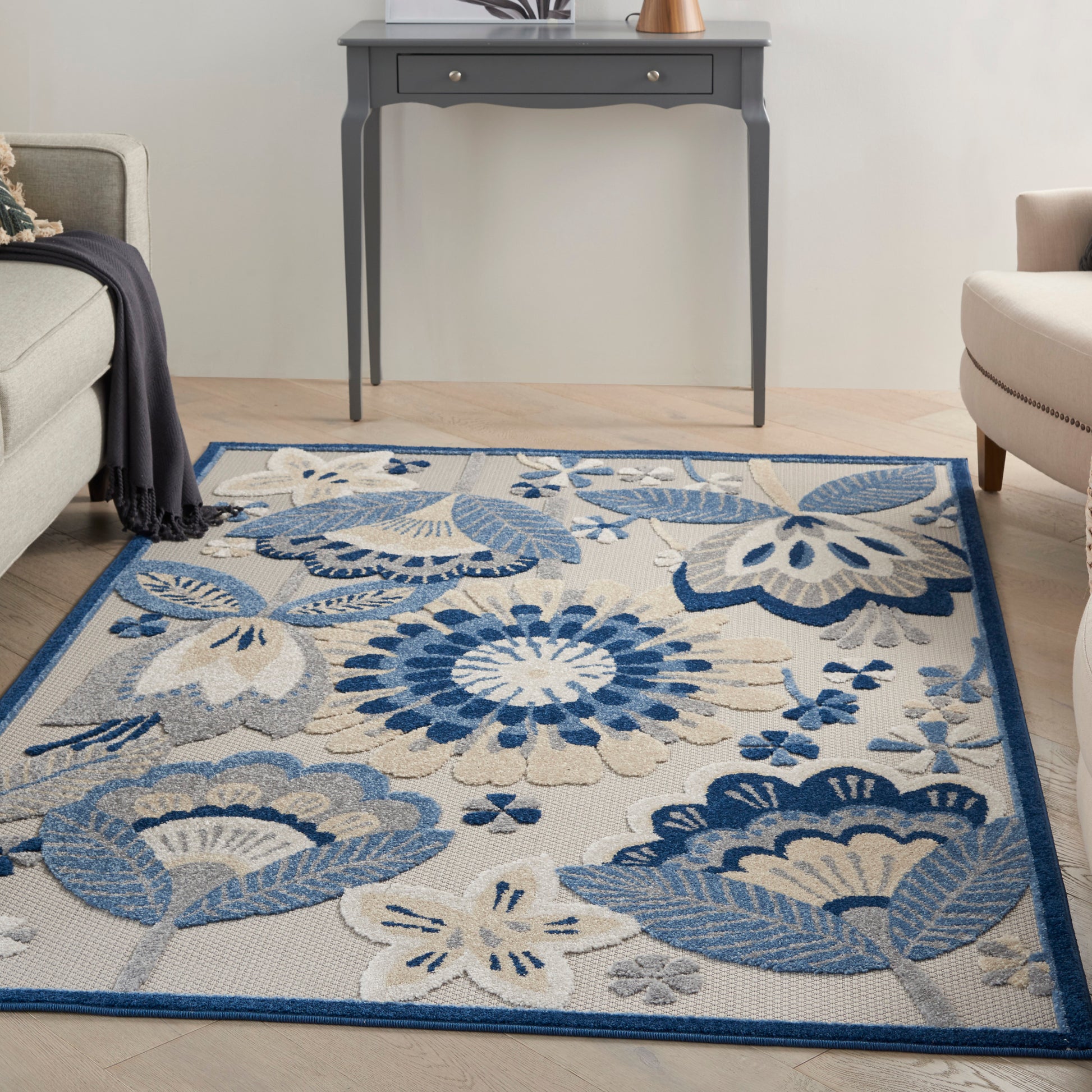 Living room with blue floral area rug on wood floor, navy border, grey console table, and beige seating.