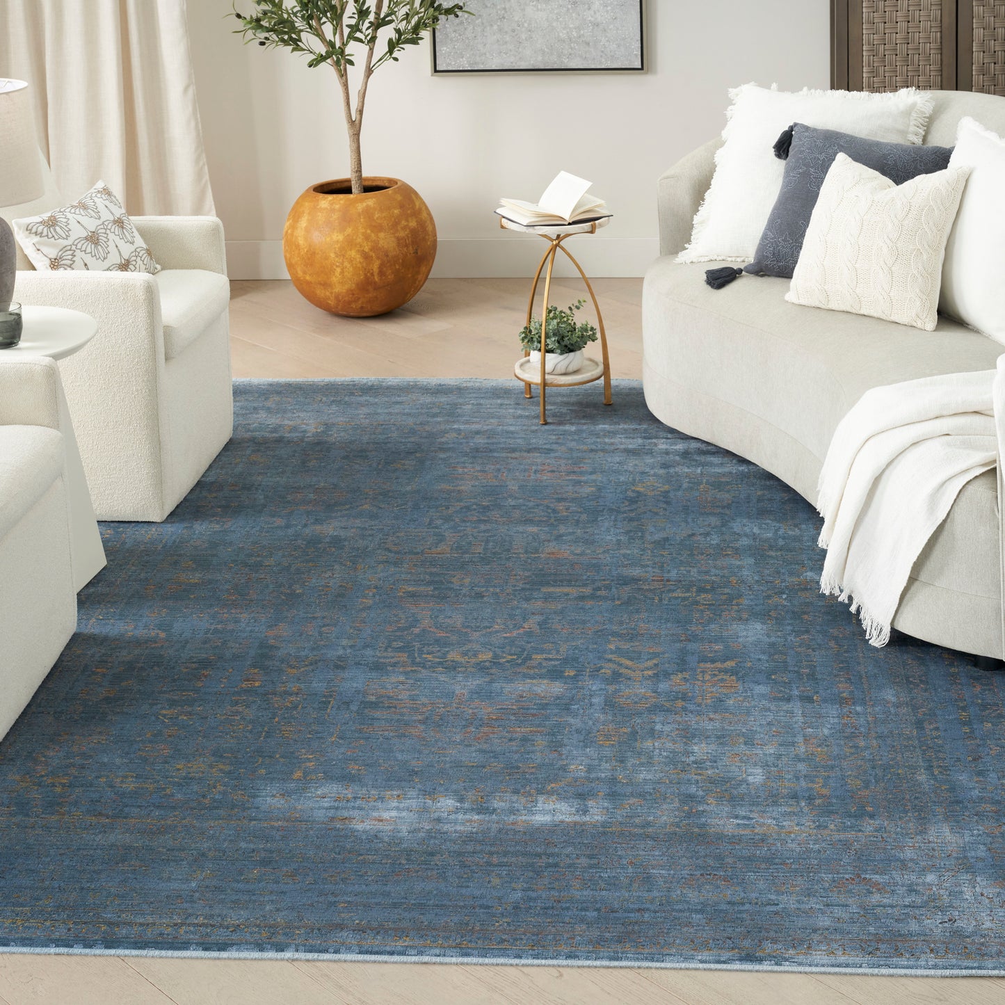 Cozy living room with a large blue rug, cream curved sofas, neutral pillows, round side table, and tall potted plant.