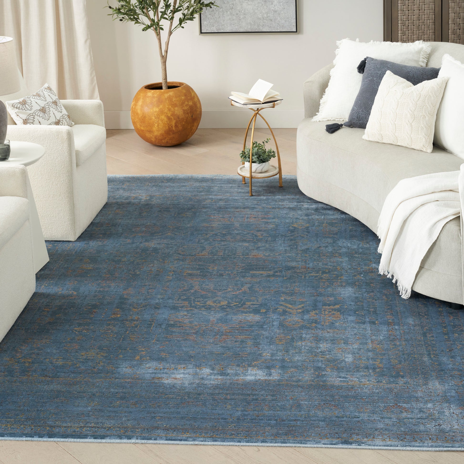 Cozy living room with a large blue rug, cream curved sofas, neutral pillows, round side table, and tall potted plant.