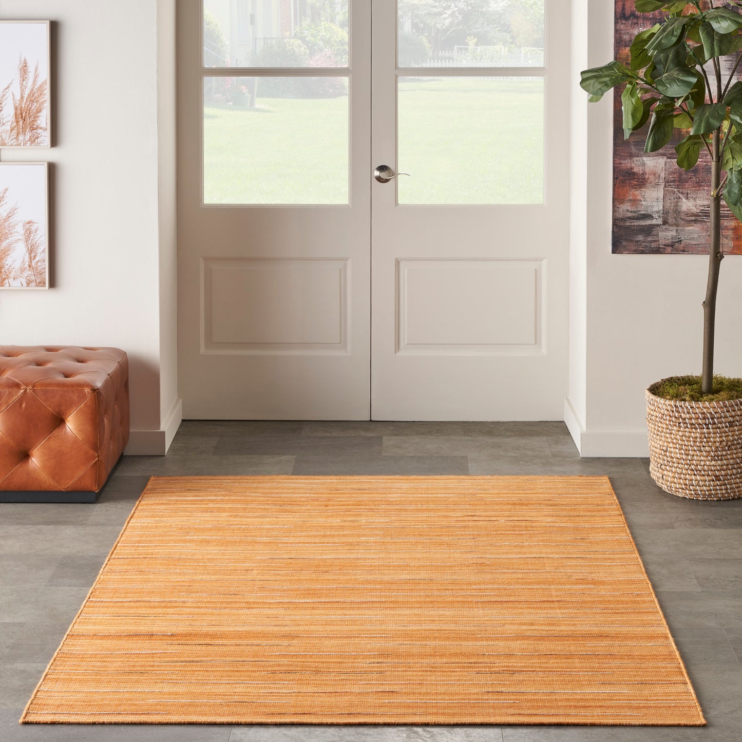 Orange rectangular striped rug in entryway with white double doors, tufted leather ottoman on left and potted plant on right.