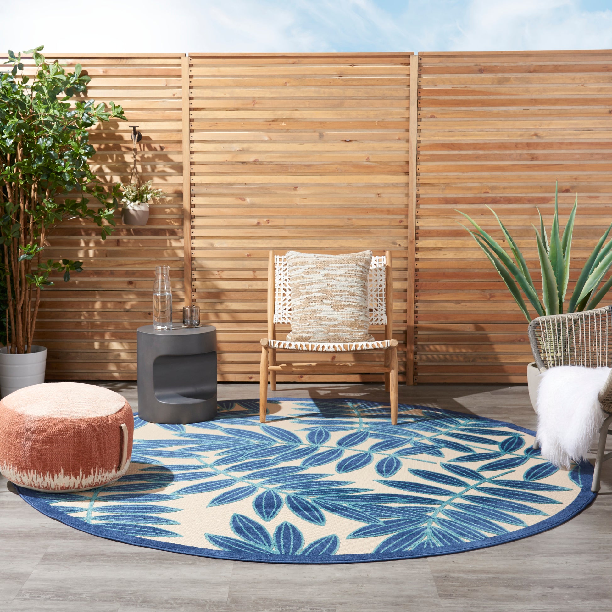 Outdoor patio with circular blue leaf rug, wood fence, rattan chair with woven cushion, peach pouf, and potted greenery.