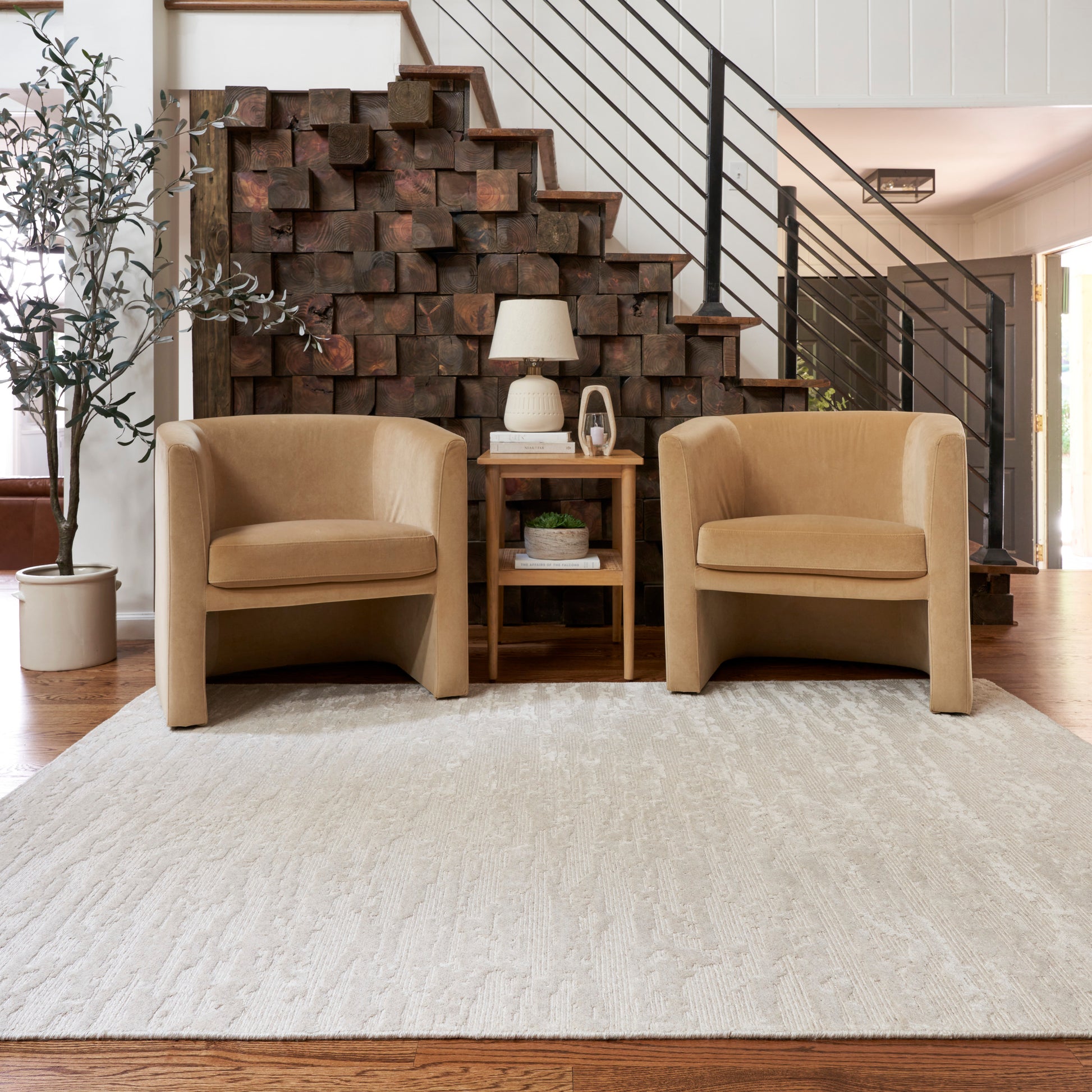 Two tan curved-arm chairs flank a small wooden side table with lamp, stacked wood staircase backdrop, beige area rug.