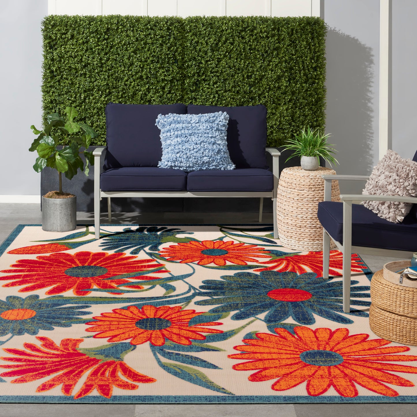 Outdoor seating area with navy sofa and chair, fluffy blue pillow, bright floral rug, potted plants and hedge wall.