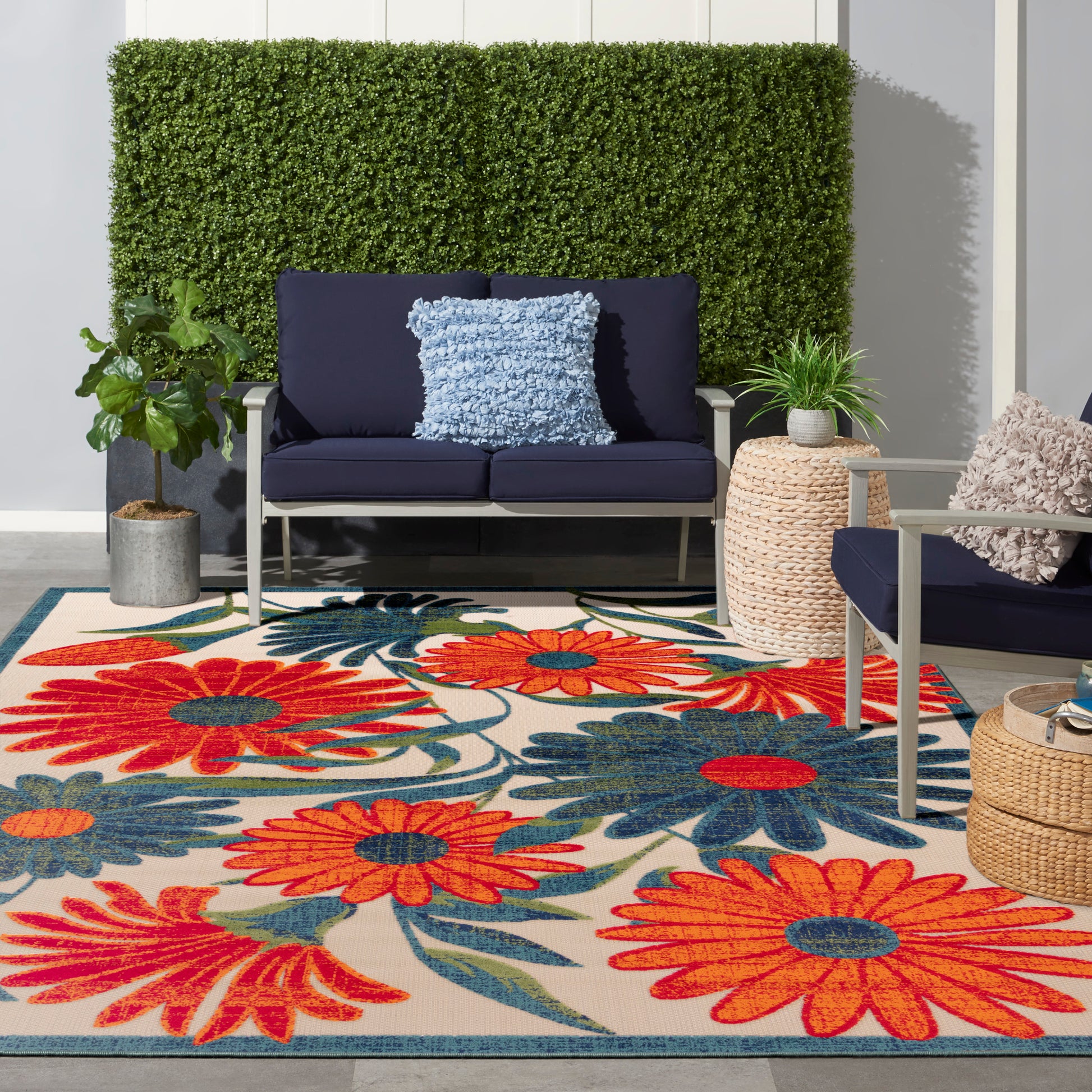 Outdoor seating area with navy sofa and chair, fluffy blue pillow, bright floral rug, potted plants and hedge wall.