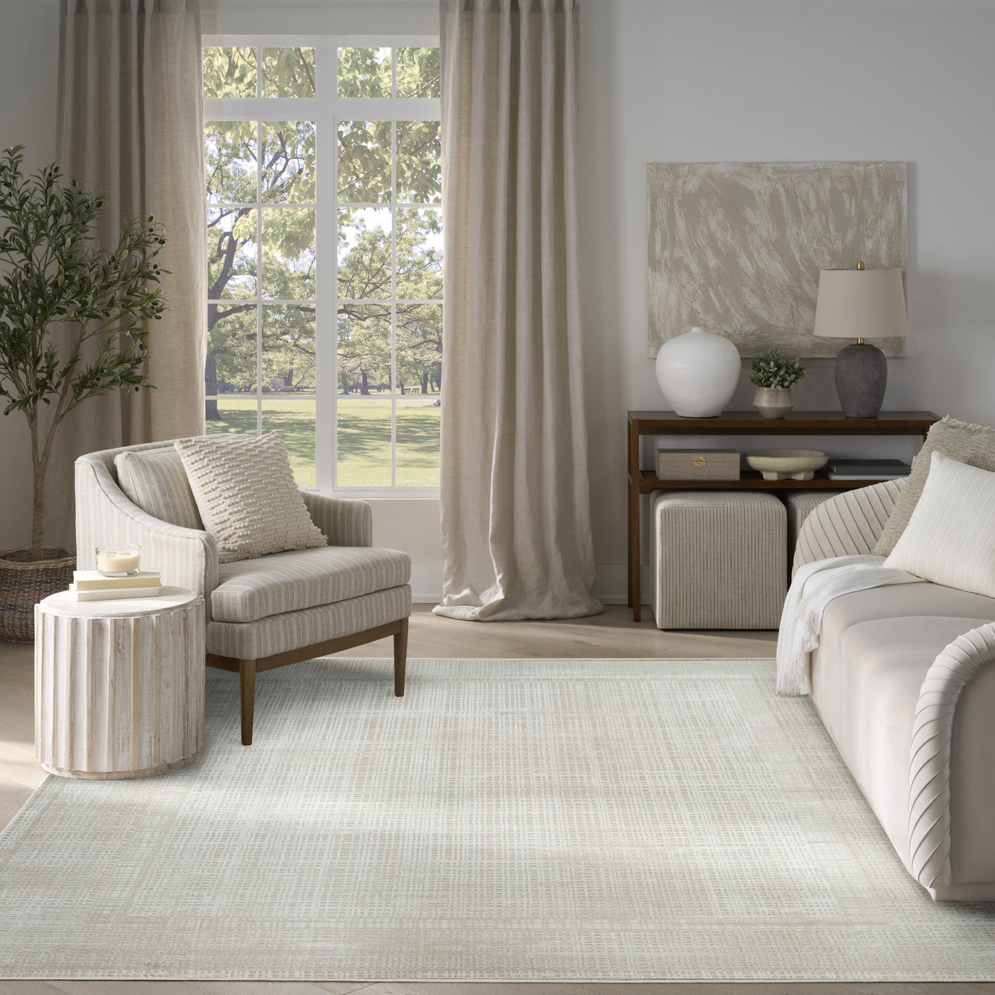 Neutral living room with beige upholstered chair & sofa, light rug, wooden side table, plant, & window with curtains.
