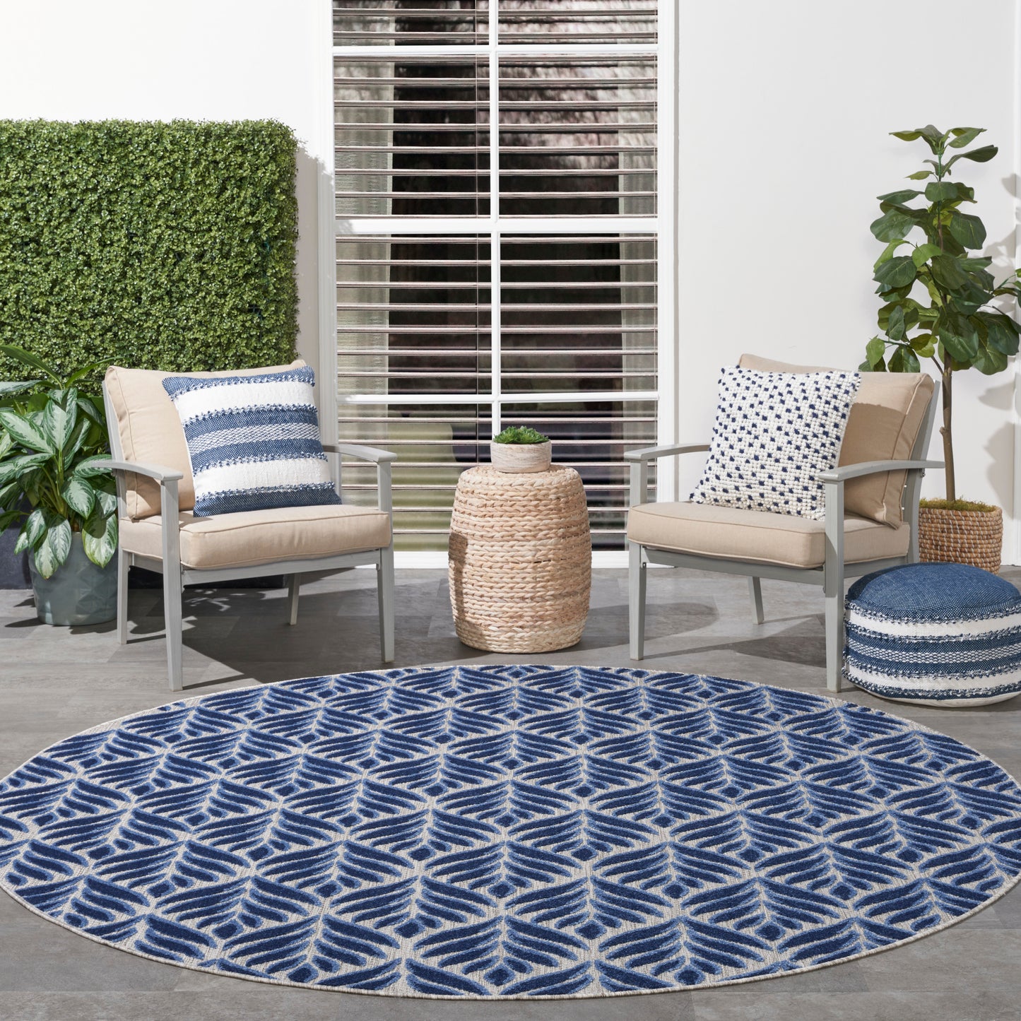 Round blue outdoor rug with white geometric leaf pattern, set on a patio between beige cushioned aluminum chairs and plants.