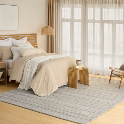 Beige linen bed with white pillows, wooden slatted headboard, rattan pendant light, light wood side table, gray striped rug, sheer curtains.