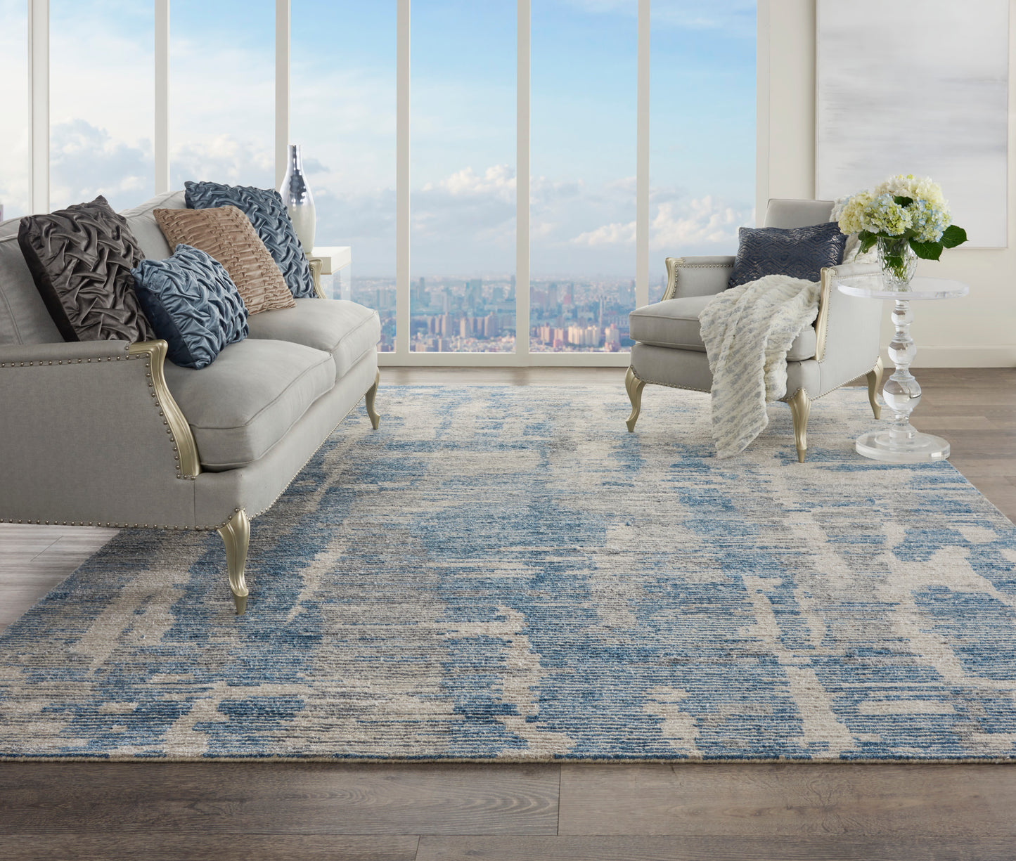 Bright living room with blue-gray patterned rug, cream sofas and armchairs, glass side table with white flowers.