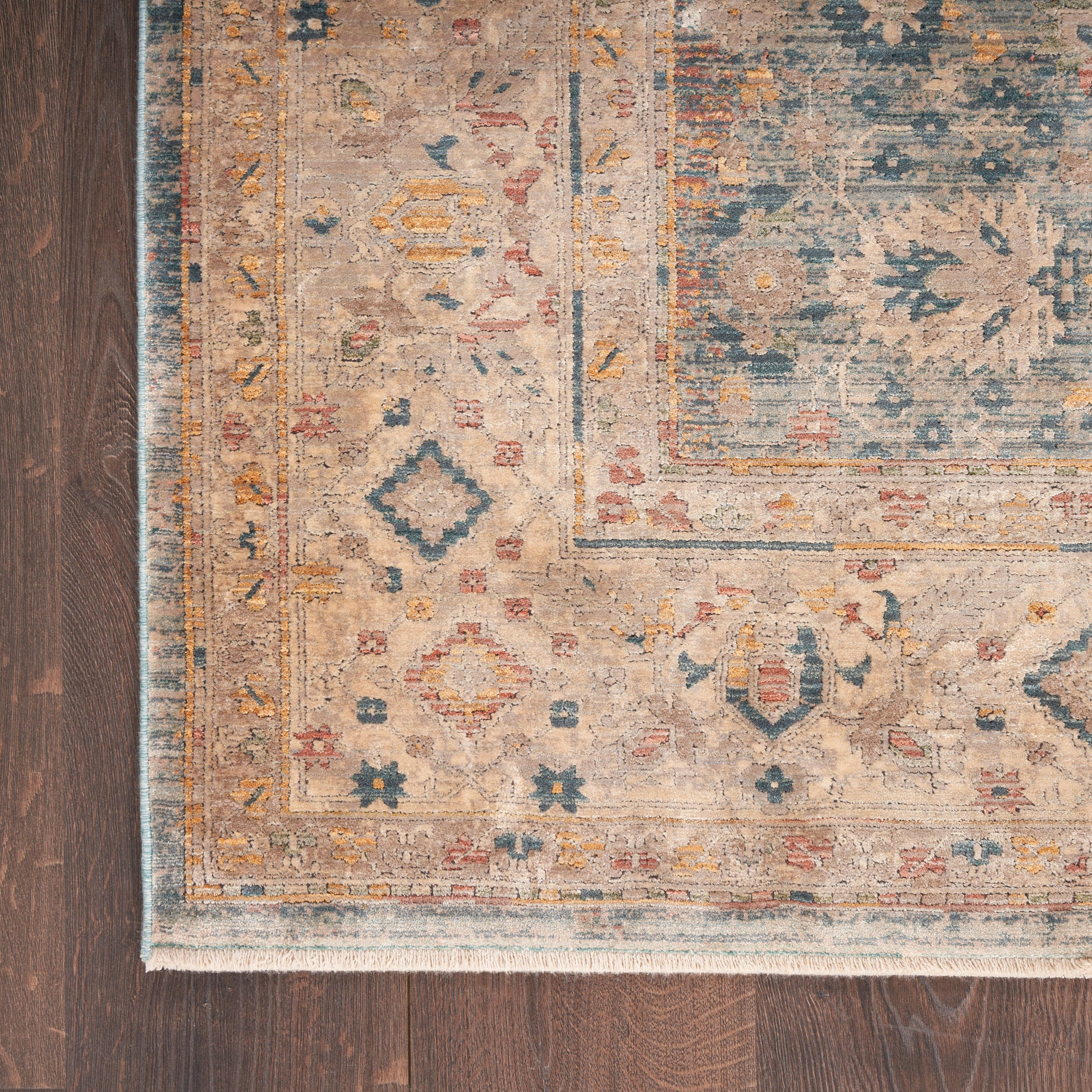 Beige oriental area rug with blue, gold and red floral-geometric border and fringed edge on dark wood floor.