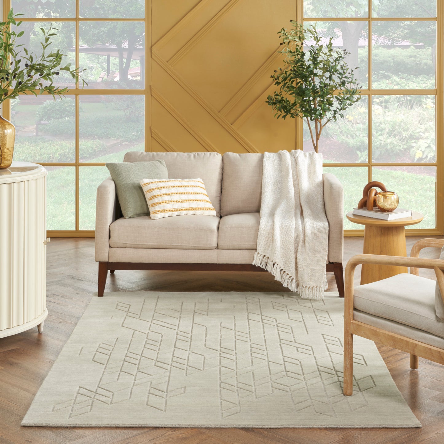 Beige sofa with light pillows and throw blanket in a bright living room; rug, wood furniture, potted plants, grid windows.