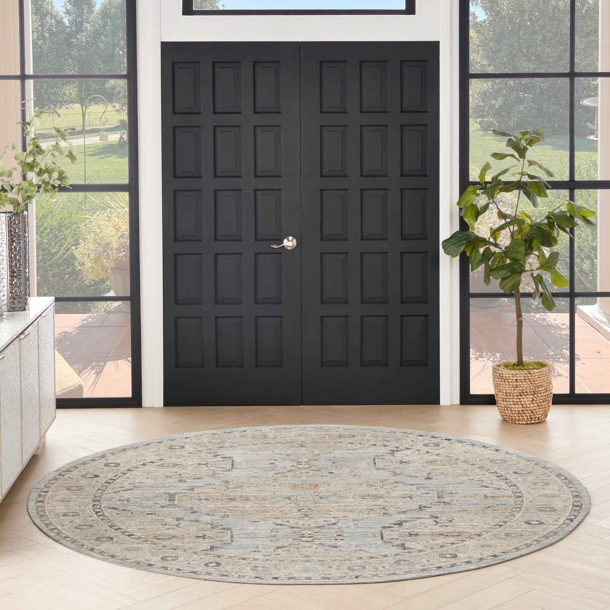 Round gray-beige patterned rug in entryway with black paneled double doors, large sidelights, & potted plant.