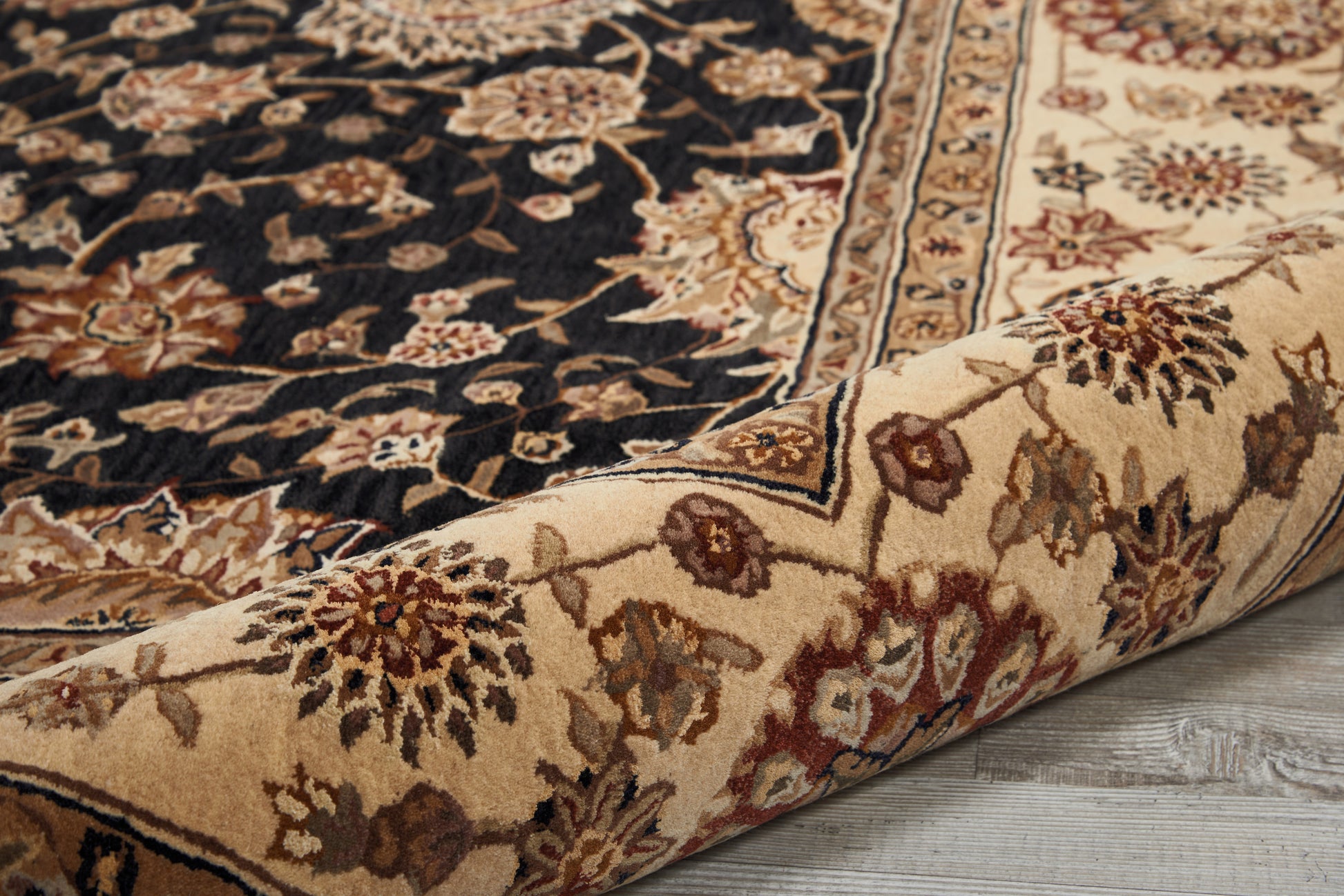 Persian-inspired area rug with a dark central field and ivory floral motifs, beige-red border, rolled on a wooden floor.