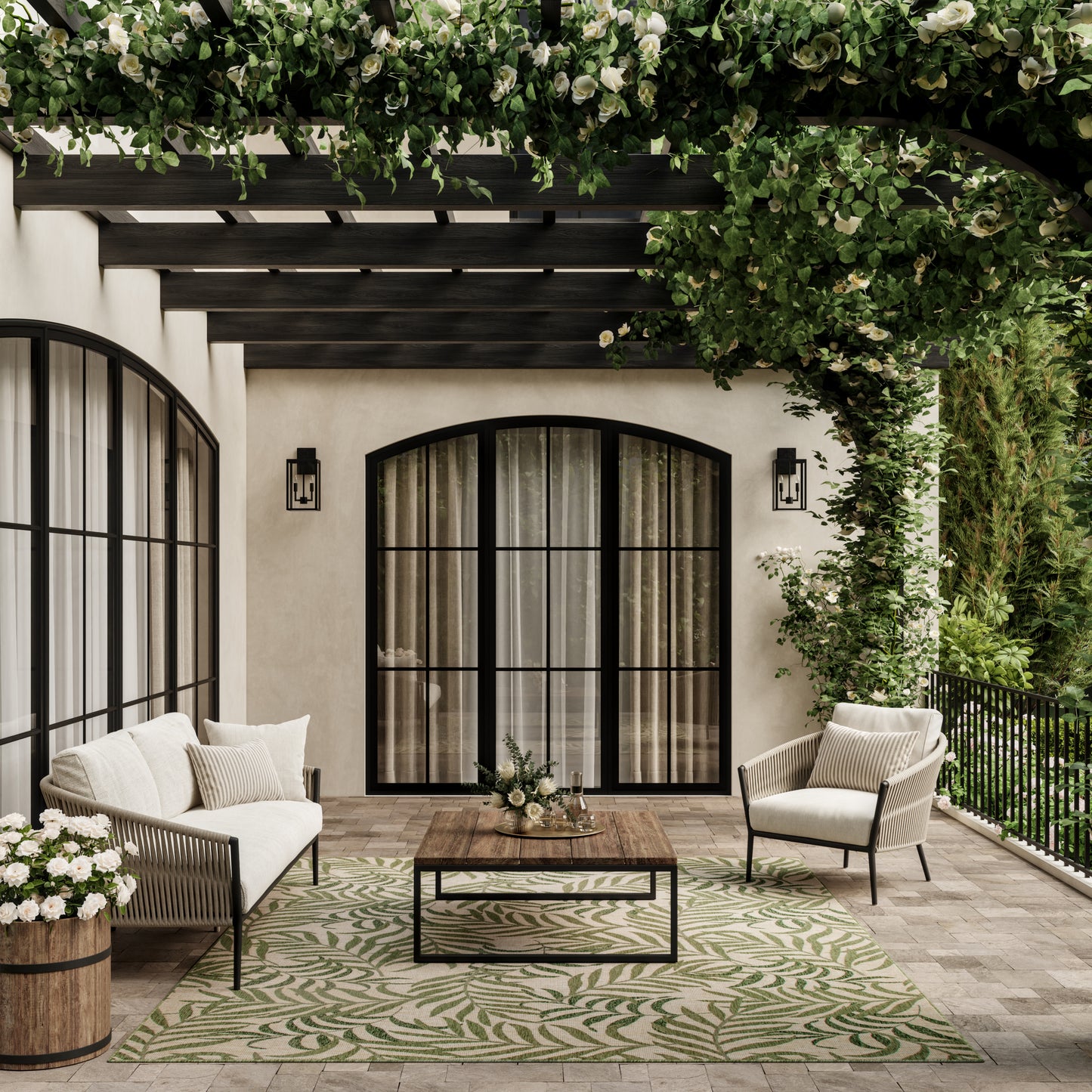 Cozy outdoor patio with arched black-framed doors, white cushions, wooden coffee table, and green rug under a leafy pergola.