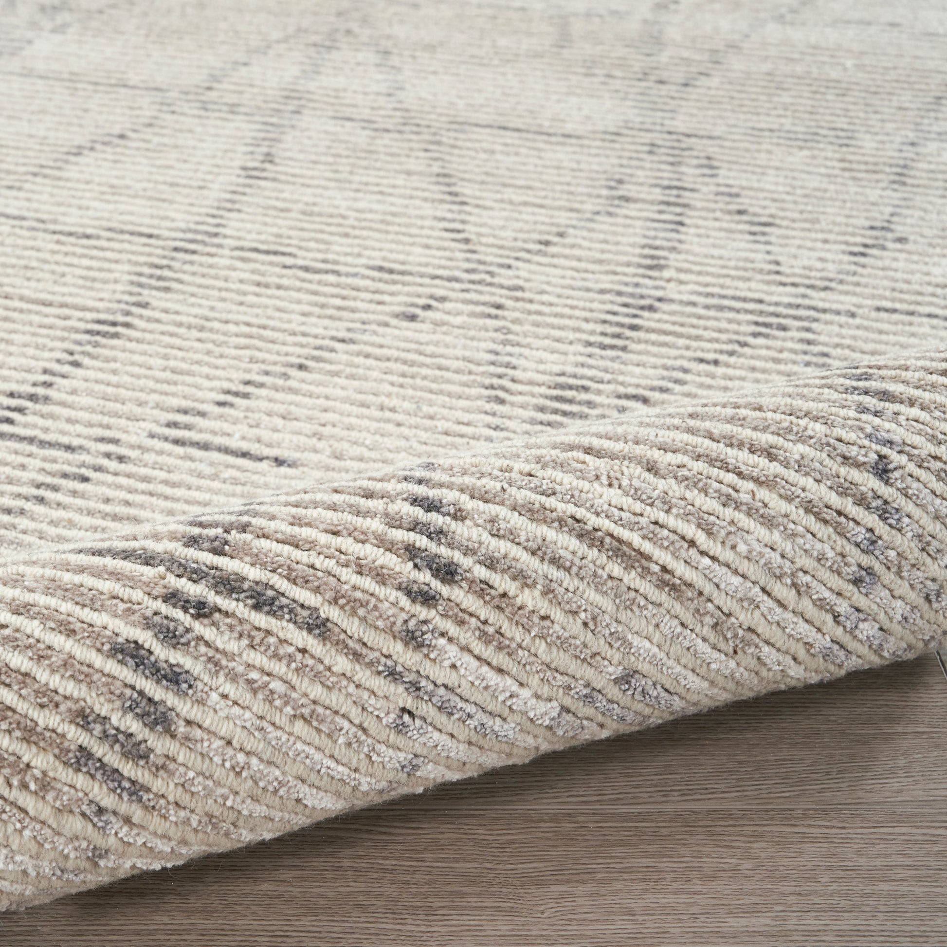 Beige textured area rug with subtle grey geometric pattern, folded edge on light wood floor.