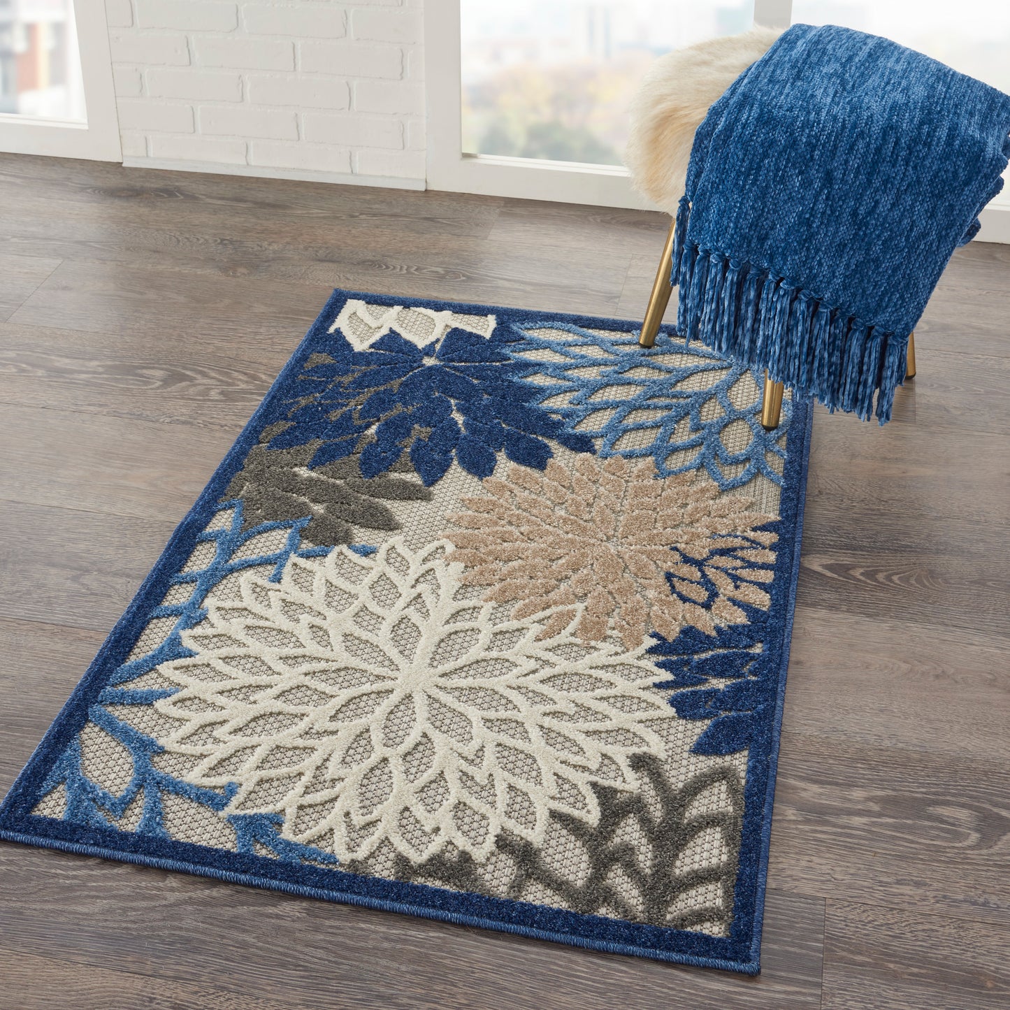 Blue-fringe area rug with large white and beige floral pattern on a dark blue border, on wooden floor beside a blue throw.