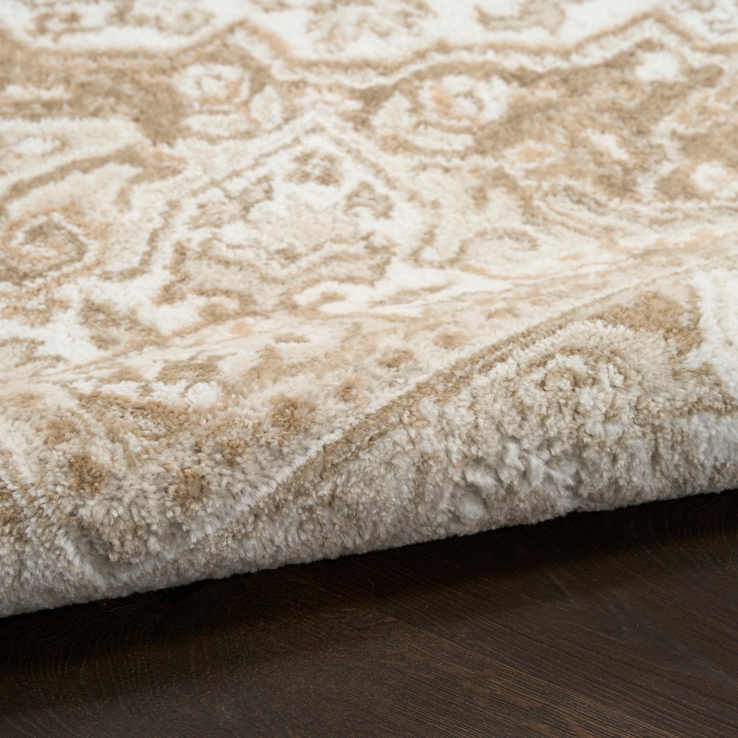 Cream-beige damask-pattern rug with thick plush pile, curled edge on dark wood floor.