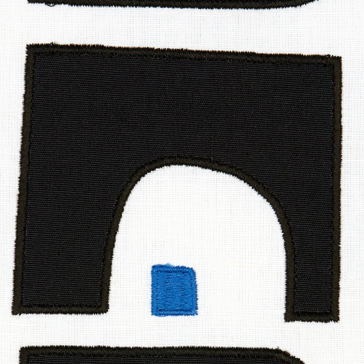Black arch-shaped embroidered patch with brown edge on white fabric; small blue square inside the arch opening.