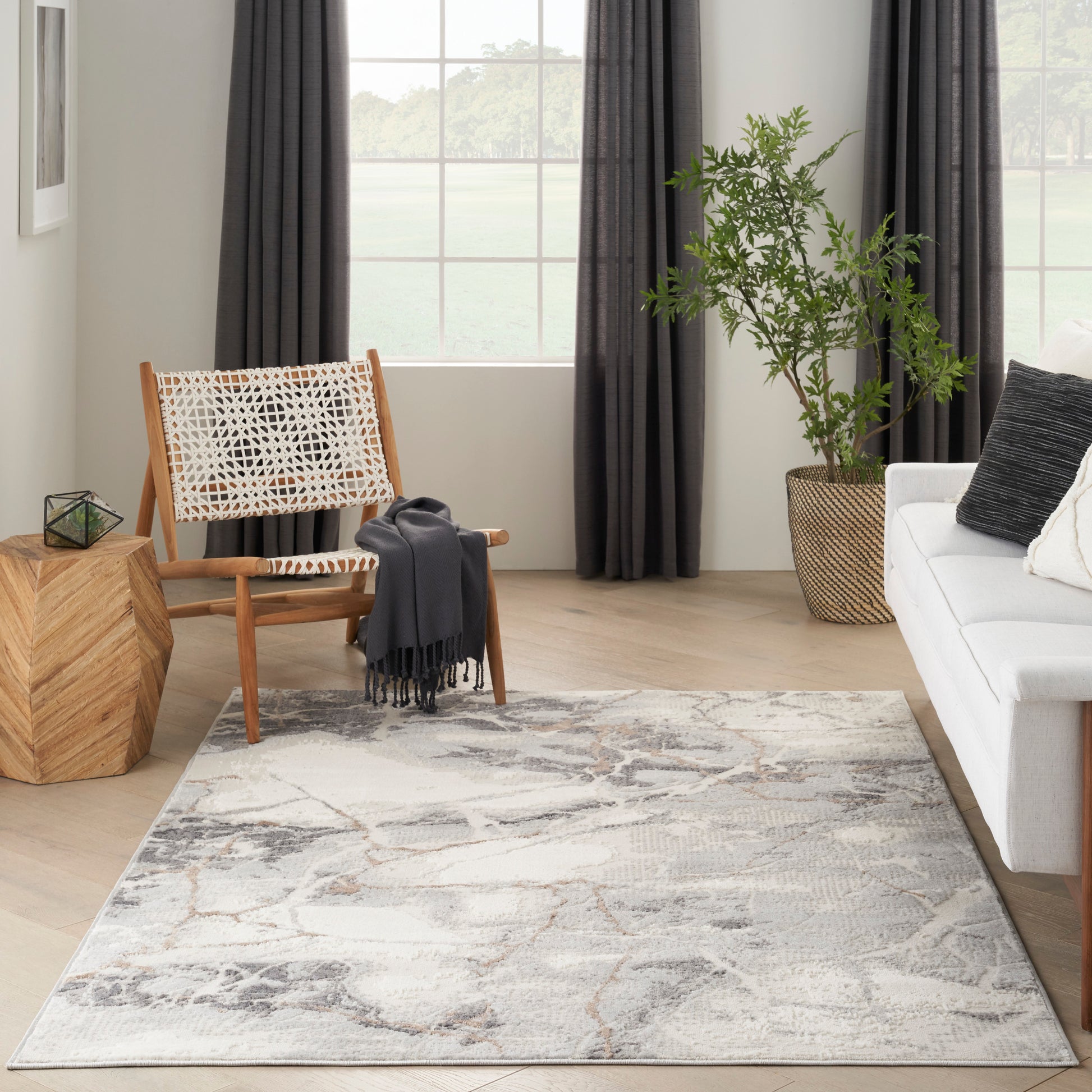 Bright living room with light wood floor, abstract gray rug, white sofa, woven wood chair, and tall plant by a large window.