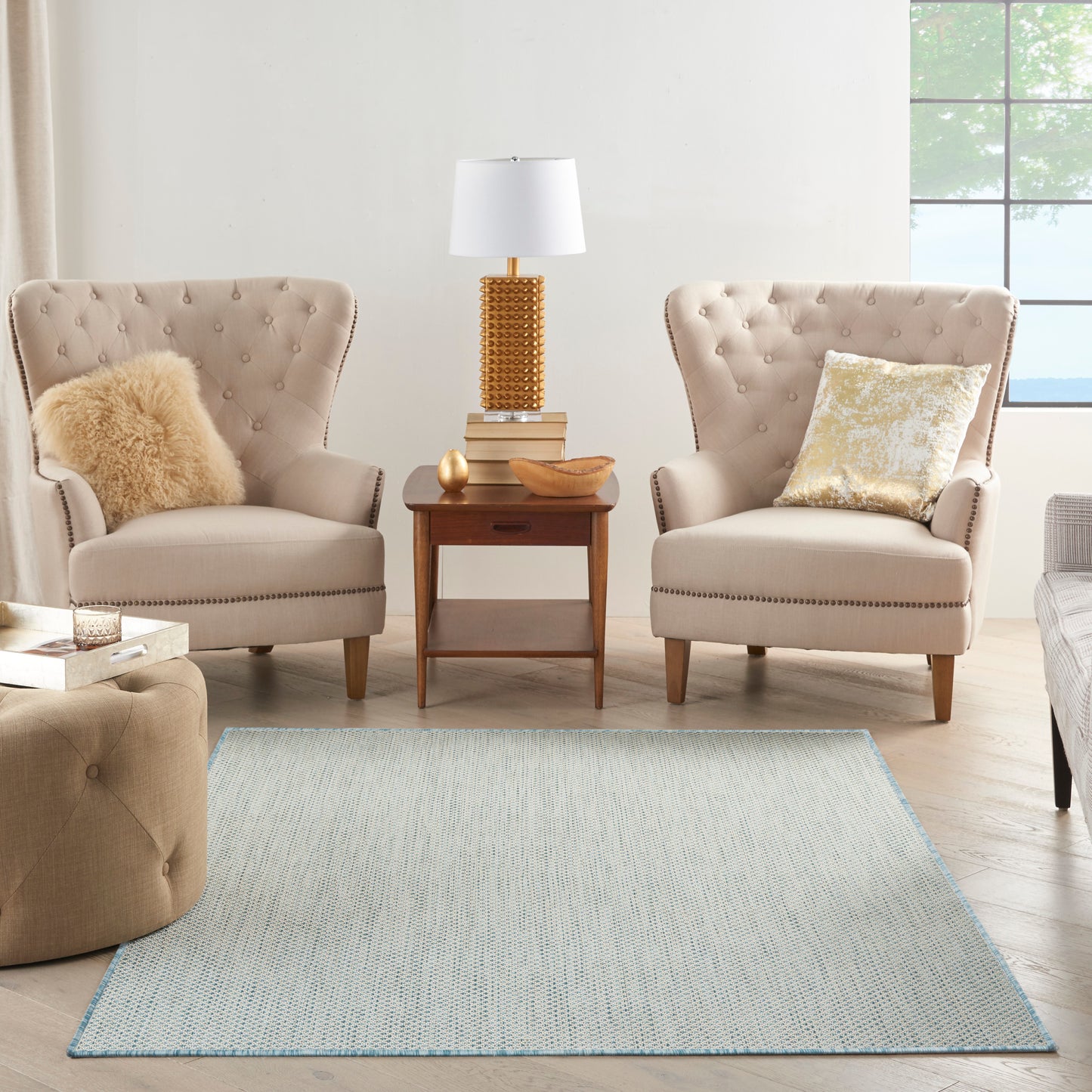 Two tufted beige armchairs with nailhead trim, wood side table, gold lamp, gold pillows, faux fur throw, light blue rug.
