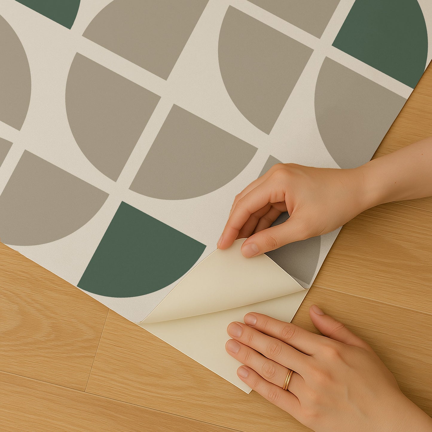 Hands peel back corner of geometric wallpaper with taupe and sage green semicircles on cream background, on wooden floor.