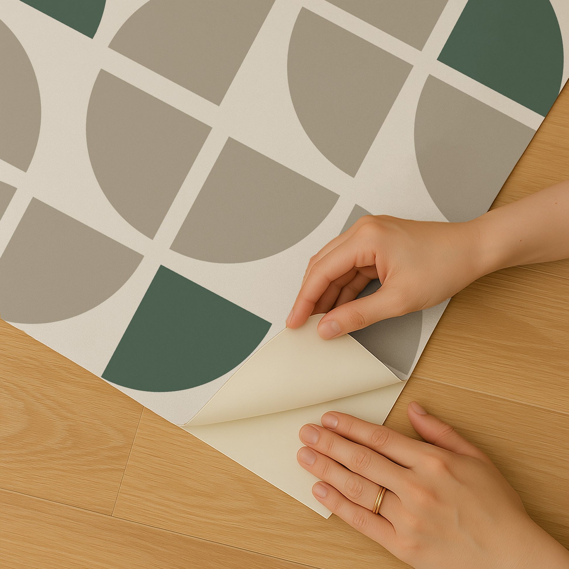 Hands peel back corner of geometric wallpaper with taupe and sage green semicircles on cream background, on wooden floor.