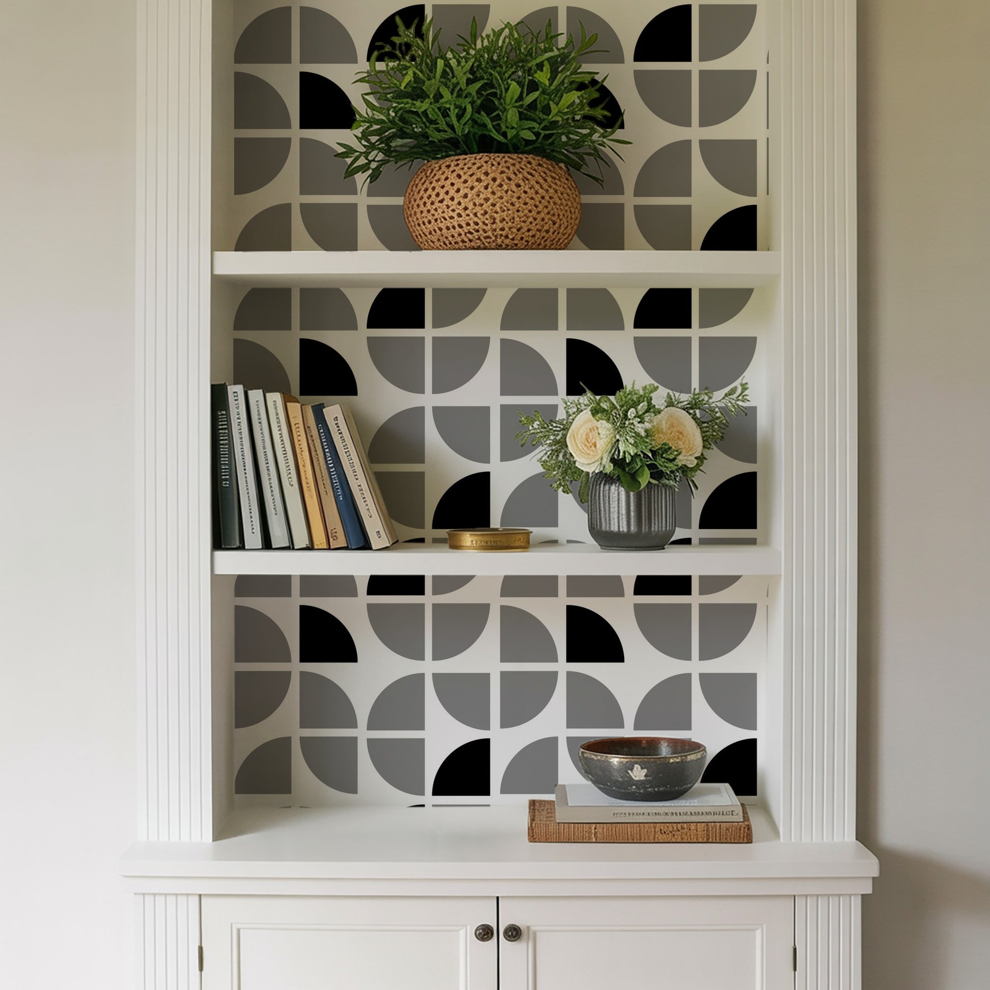 White built-in shelf with gray geometric wallpaper; top wicker plant, middle books and gray vase with cream flowers, lower bowl on books.