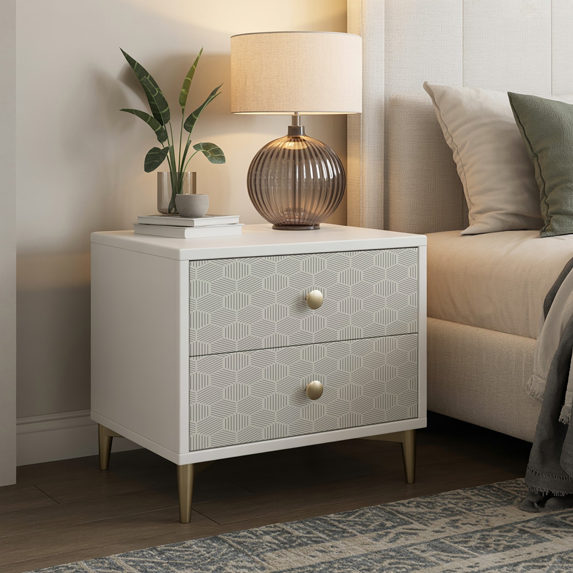 White two-drawer nightstand with geometric patterned fronts, brass knobs, tapered gold legs; lamp, plant, and books on top.