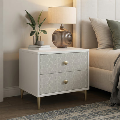 White two-drawer nightstand with geometric patterned fronts, brass knobs, tapered gold legs; lamp, plant, and books on top.