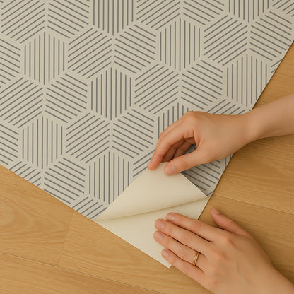 Gray peel-and-stick wallpaper with a geometric hexagonal mosaic pattern being peeled back by hands over a wooden floor.