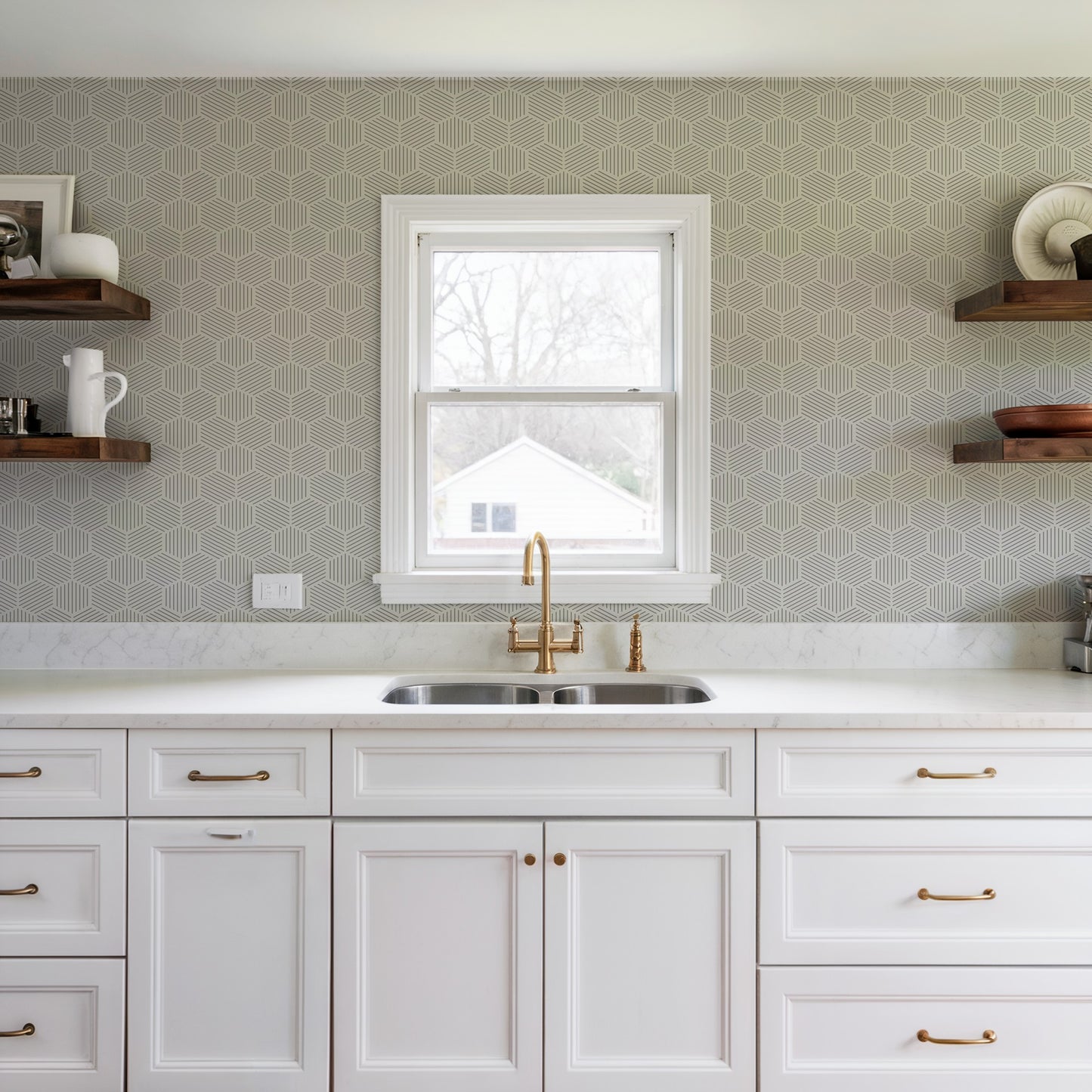 White kitchen with marble countertop, brass faucet and stainless double sink under a window, featuring striped hexagonal mosaic gray peel-and-stick wallpaper.