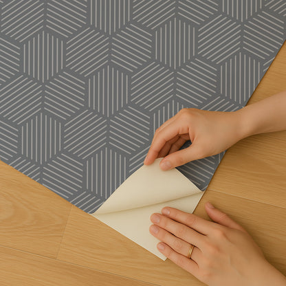 Blue peel-and-stick wallpaper with a striped hexagonal mosaic pattern being peeled back by hands on a wooden floor