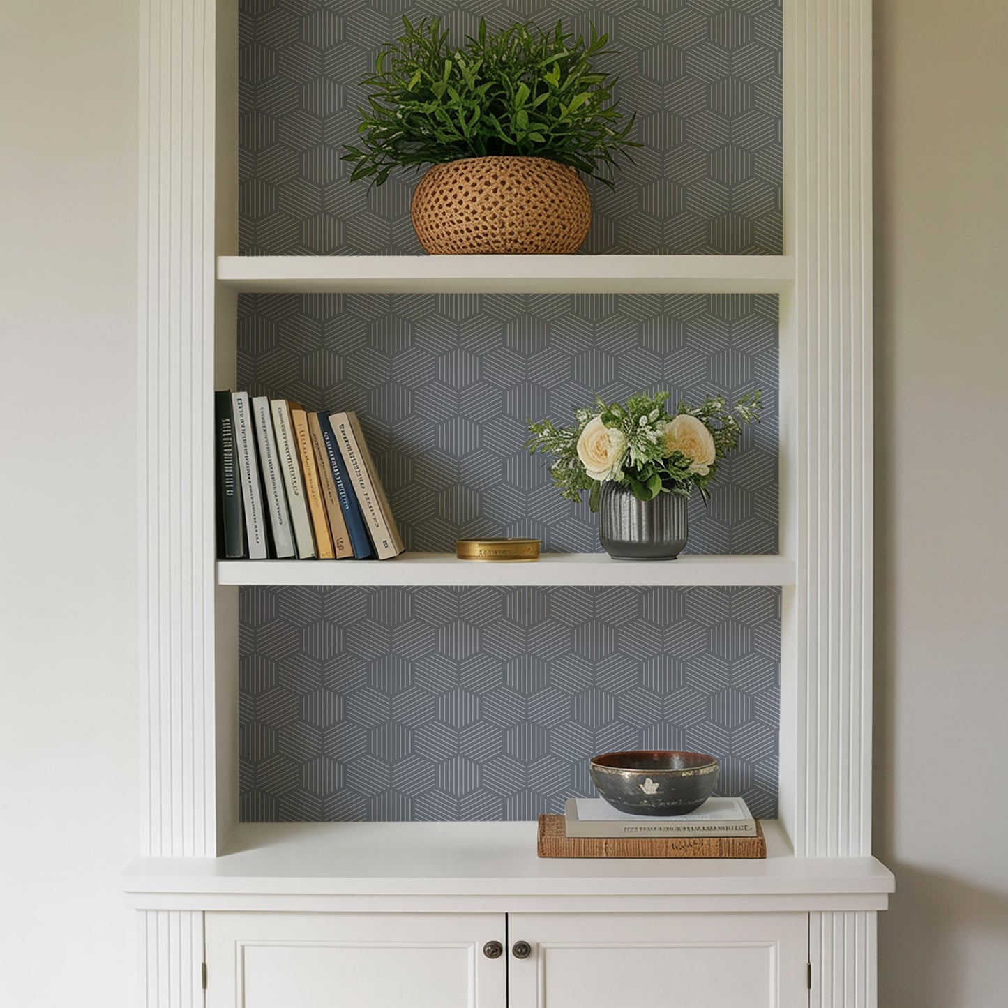 White built-in bookcase with geometric gray wallpaper; top woven plant basket, middle books and cream rose vase, bottom bowl on books.