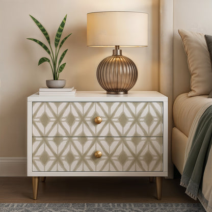 White two-drawer nightstand with gold knobs, geometric beige pattern; ribbed bronze table lamp, plant, and books on top.